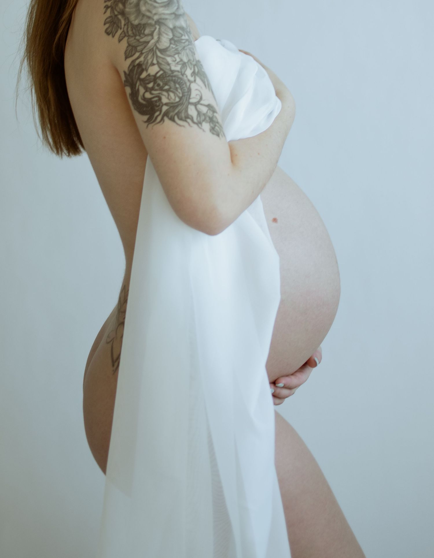 Artistic maternity photos showing a woman in white against a light background with visible tattoos.
