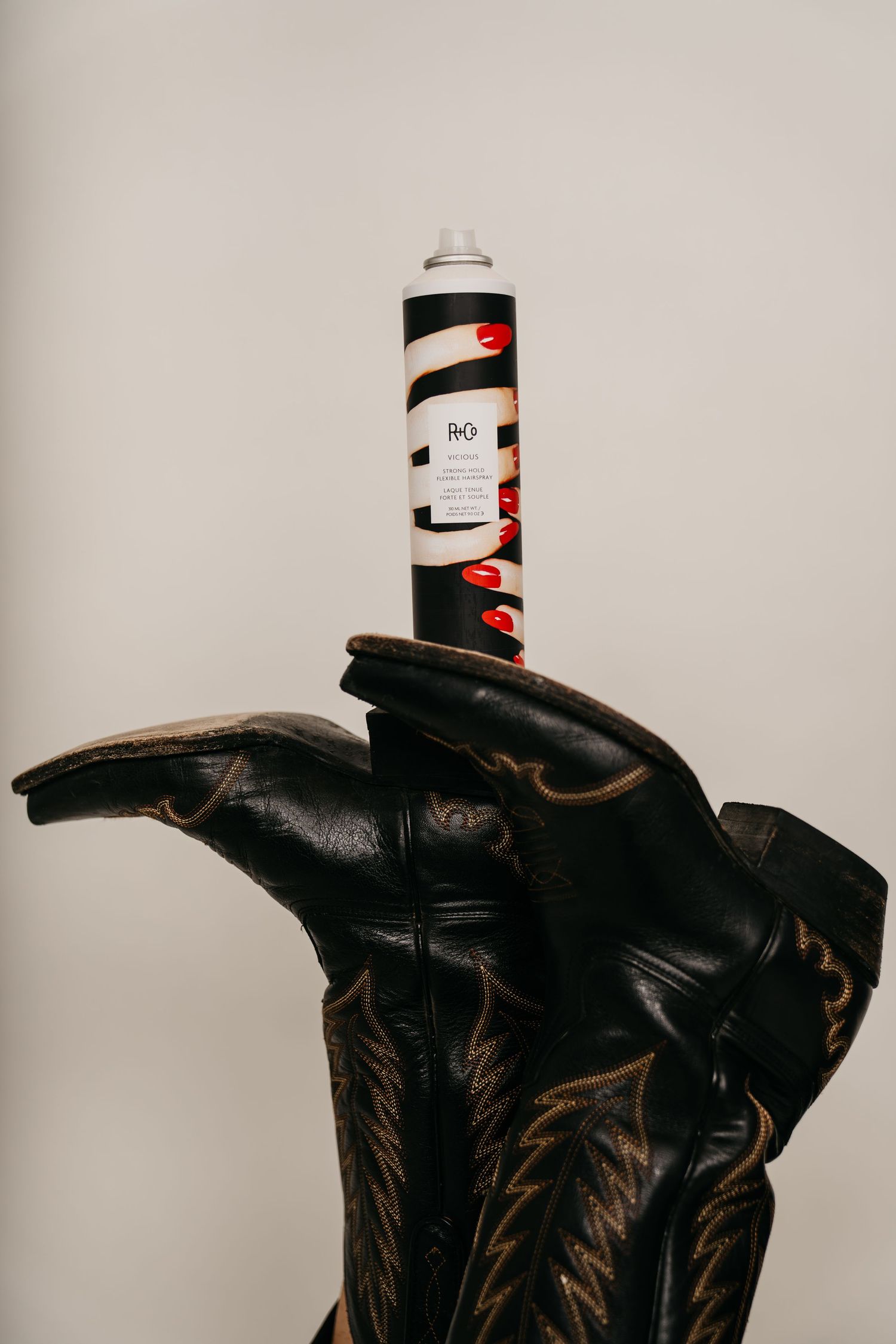 R+Co hair spray balanced on black leather cowboy boots against a neutral gray background.