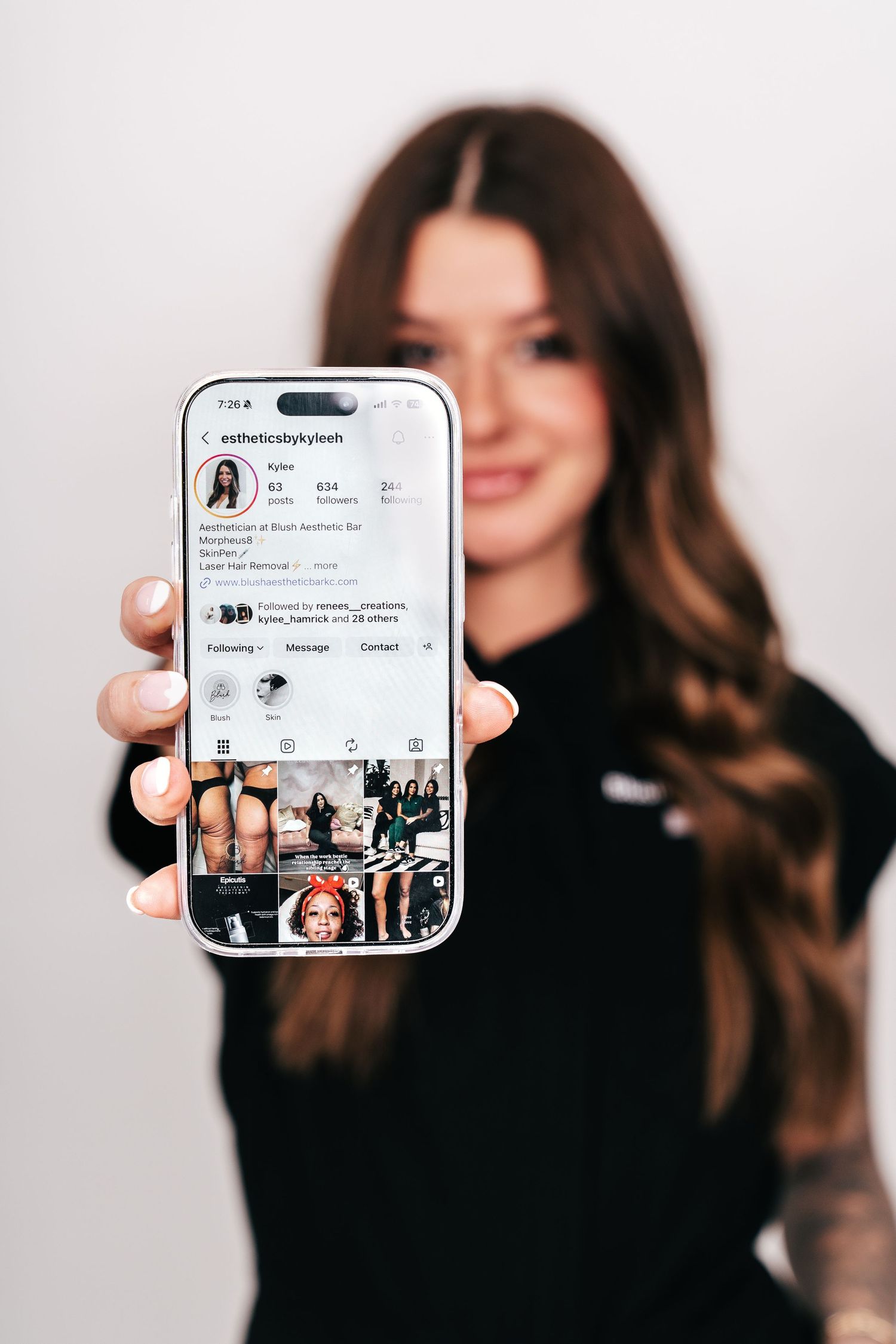 Young woman in black top holding smartphone displaying Instagram profile page with grid of photos.