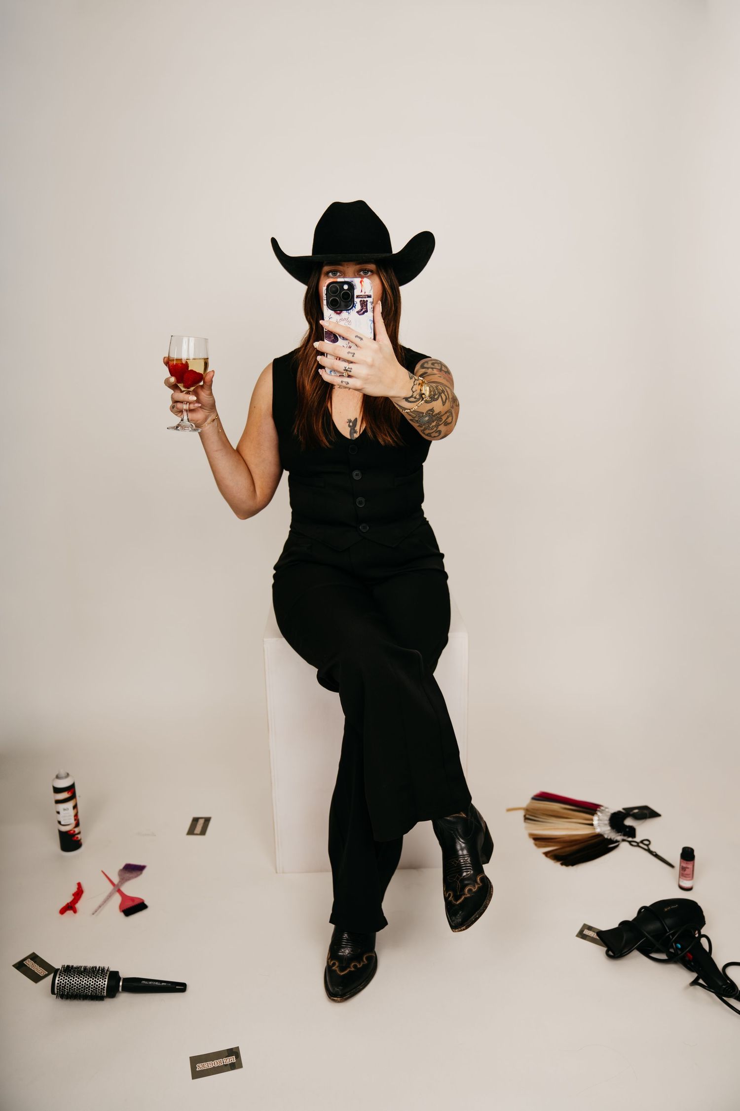 Woman in black cowboy hat and outfit holds wine glass while taking selfie, surrounded by hair tools on white background.