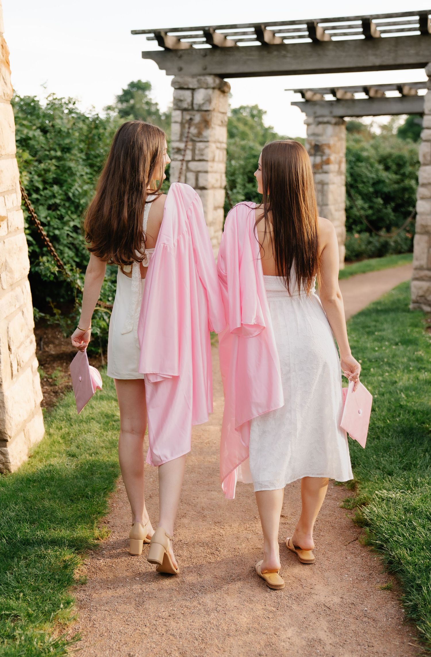 Two women in white dresses carrying pink wraps walk along a garden path beneath a stone pergola.