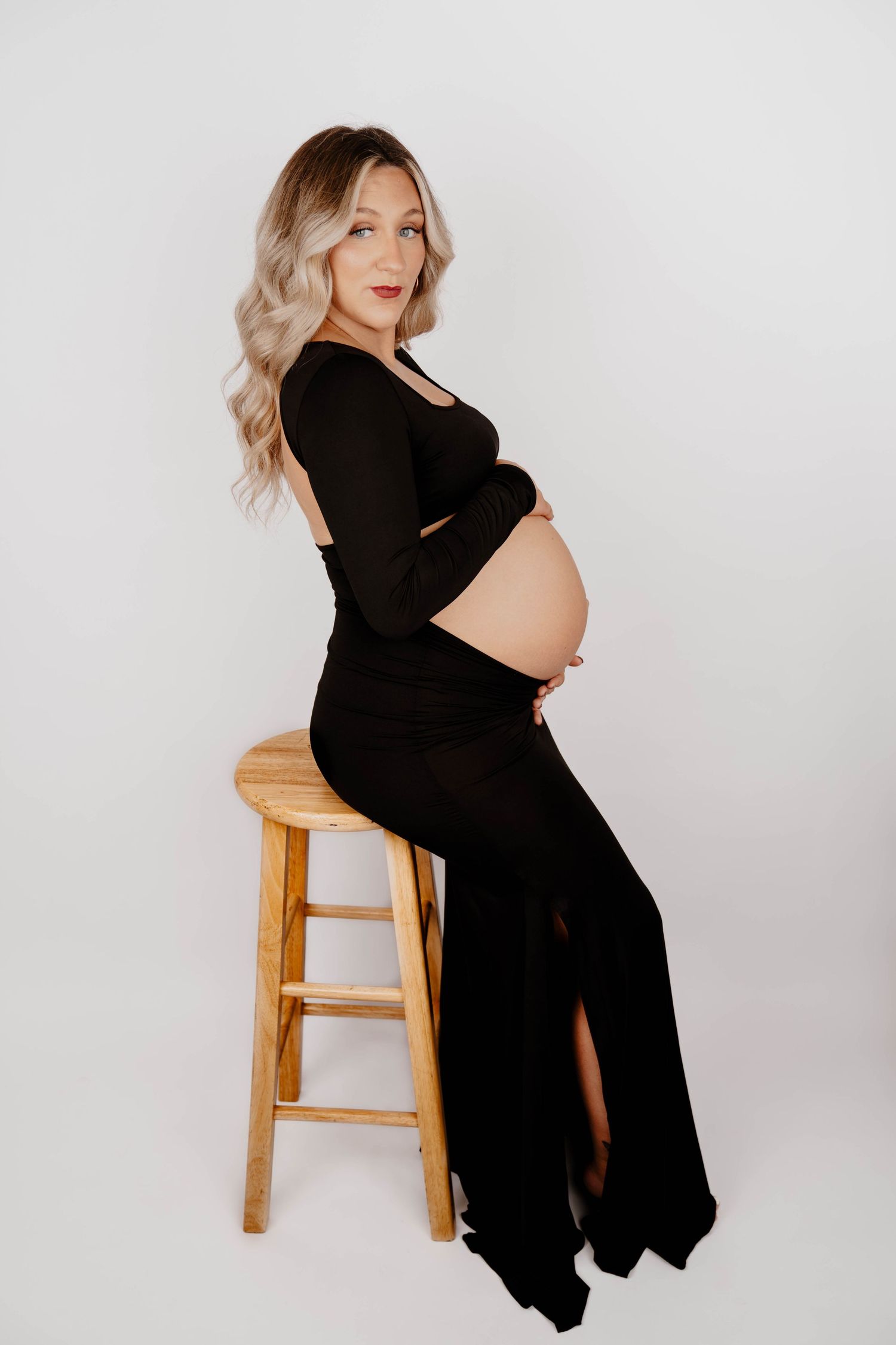 An expectant mother poses on a wooden stool in a black formal dress.