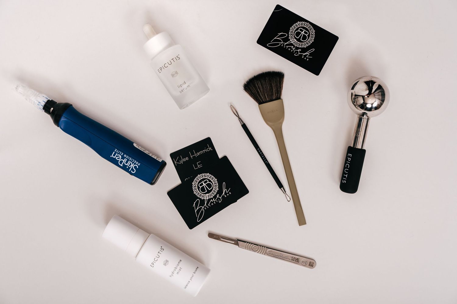 Flat lay of beauty tools including brushes, tweezers, serums, and makeup accessories on a white background.
