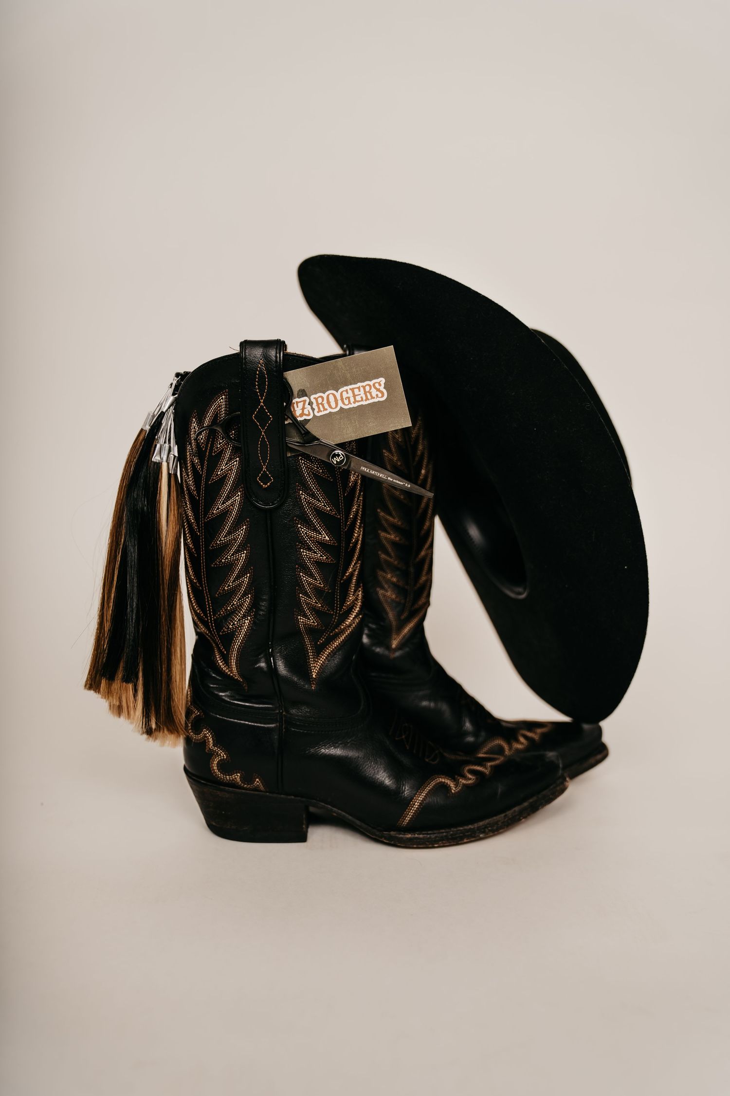 Black cowboy boots with gold embroidery, tassels, and a black cowboy hat on a neutral background.