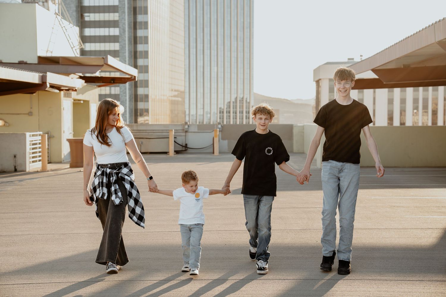Family of four holding hands walking on rooftop plaza with city buildings in background on sunny day.