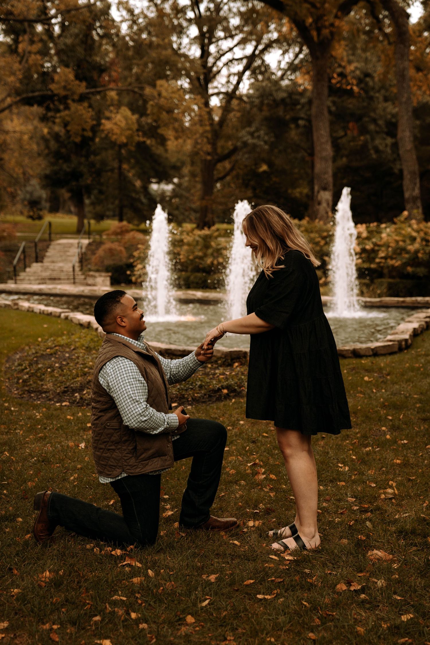 A romantic proposal scene in front of fountains with fall foliage in the background.