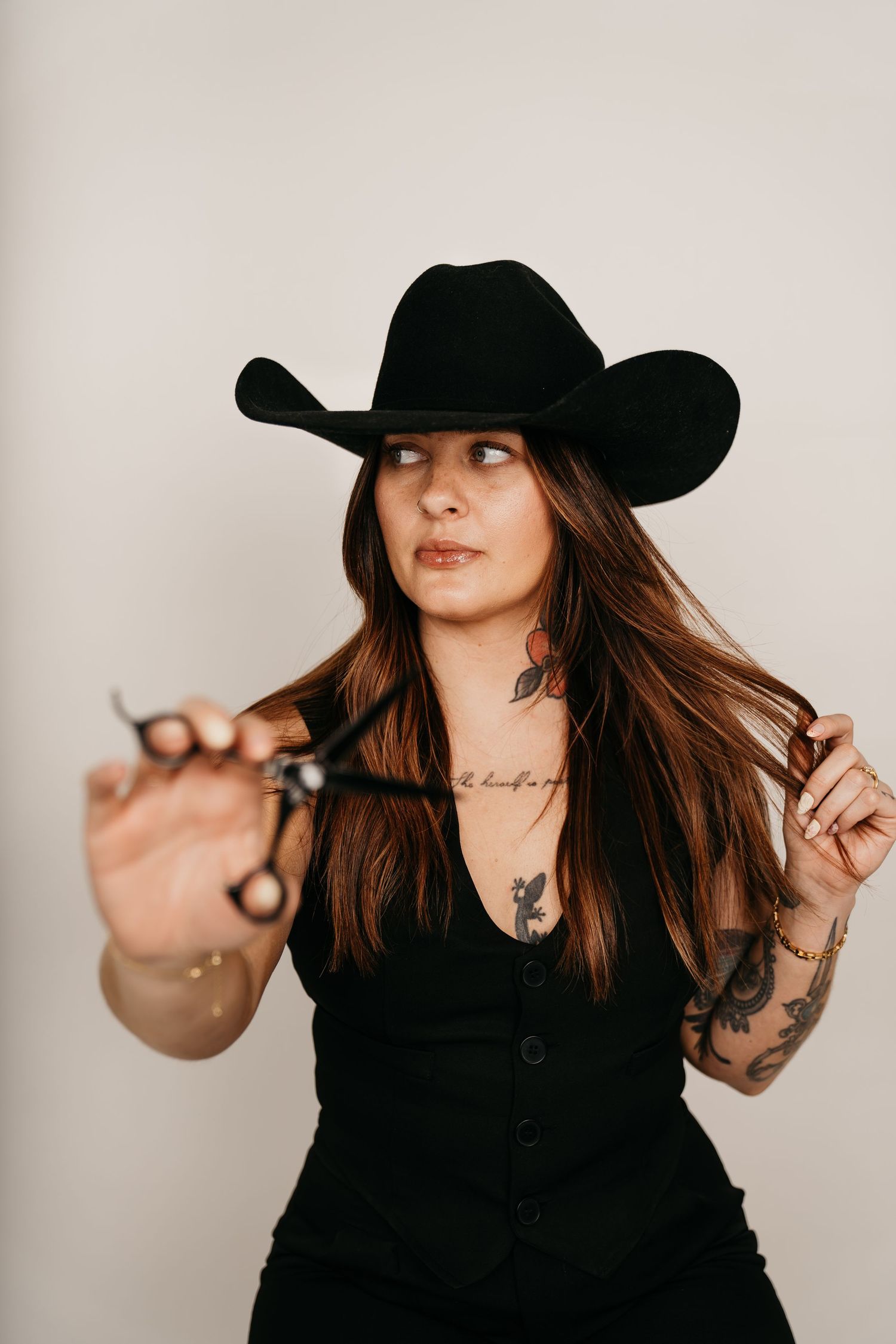 Tattooed woman in black cowboy hat and outfit holds scissors, posing against light gray background.