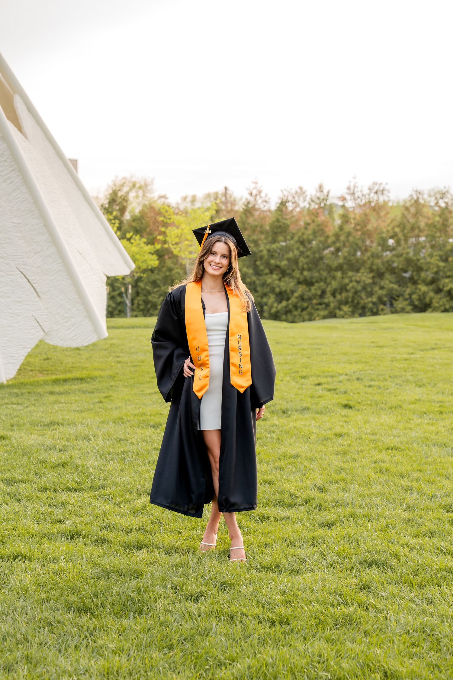 Young woman in black graduation gown and cap with gold stole smiling outdoors on green grass.