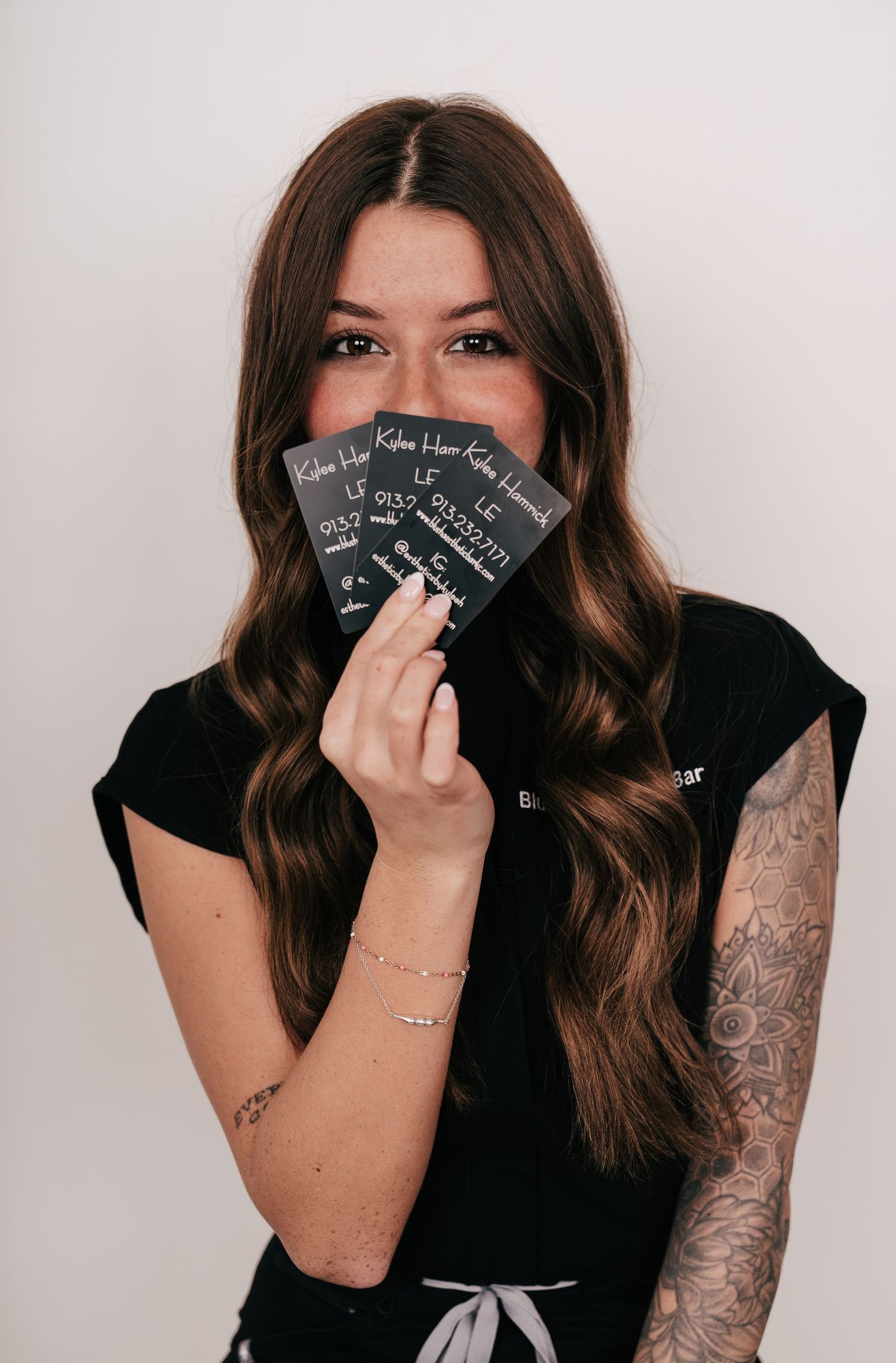 Tattooed woman with long brown hair holding dark business cards in front of her face, wearing black outfit.