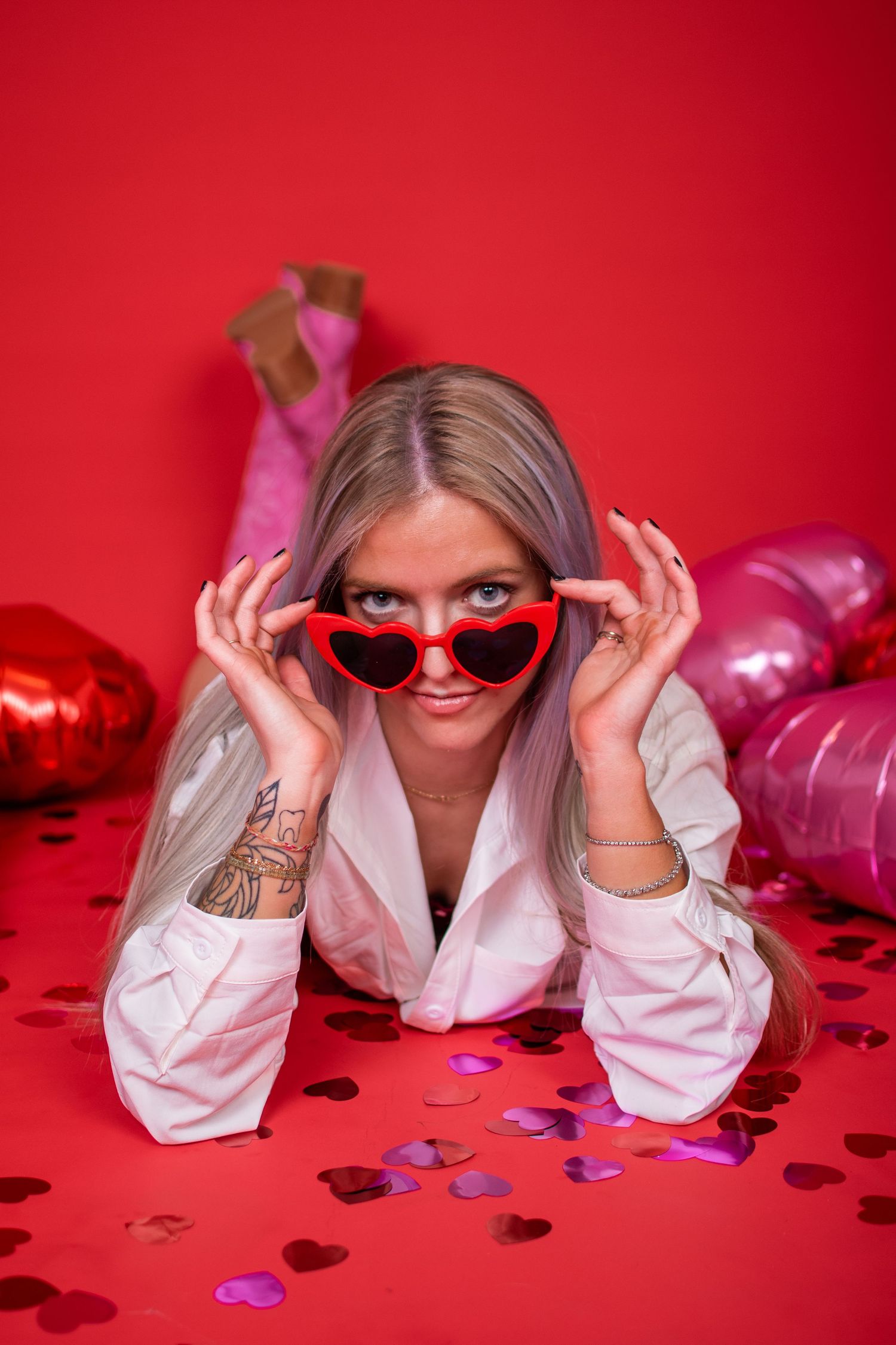 Fun portrait series with red heart sunglasses and confetti on vibrant red background.
