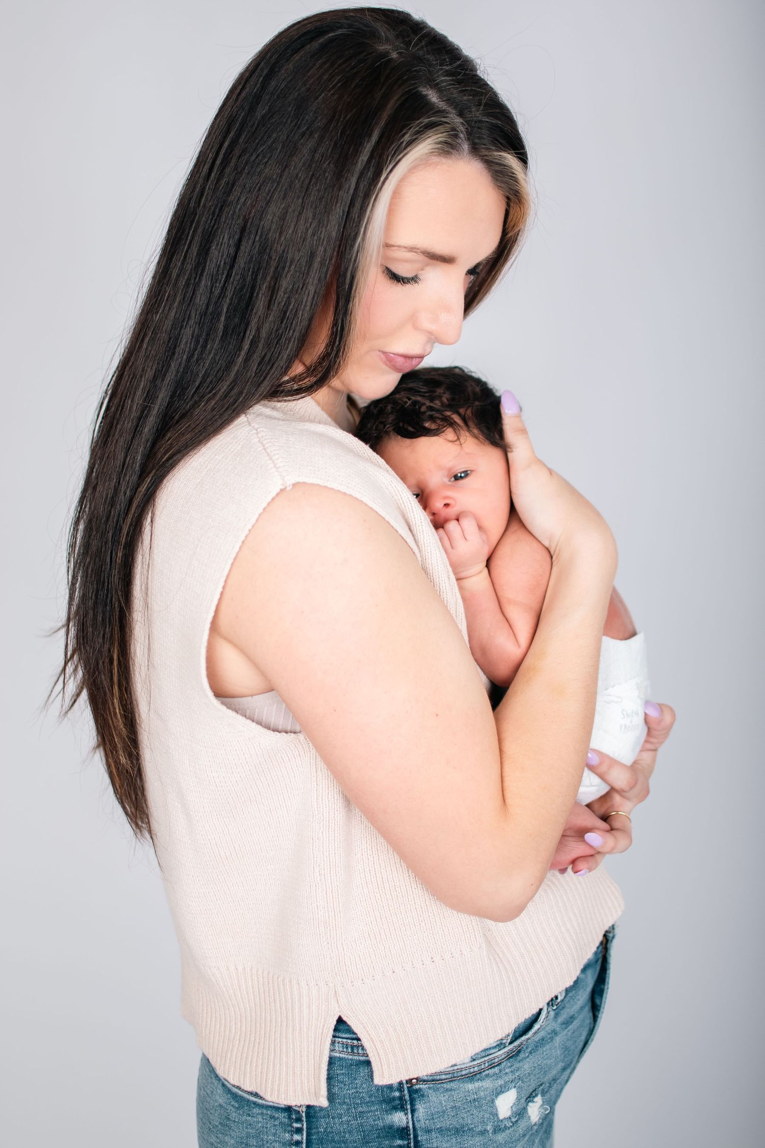 Mother in cream sweater holding sleeping newborn baby against white background.