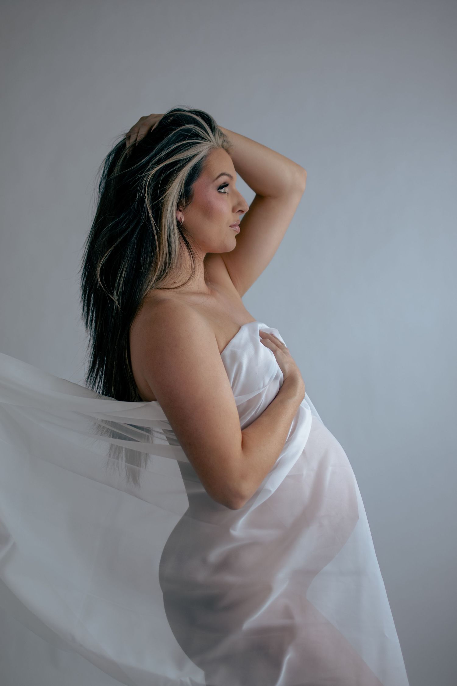 Ethereal maternity portrait with sheer white fabric in dramatic lighting.