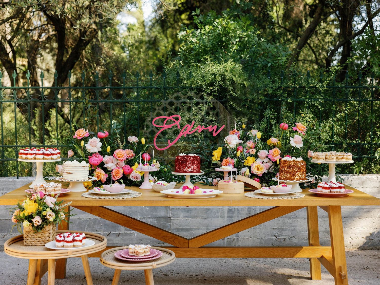 A rustic outdoor dessert table decorated with pink and white flowers, cakes, and treats in a garden setting.