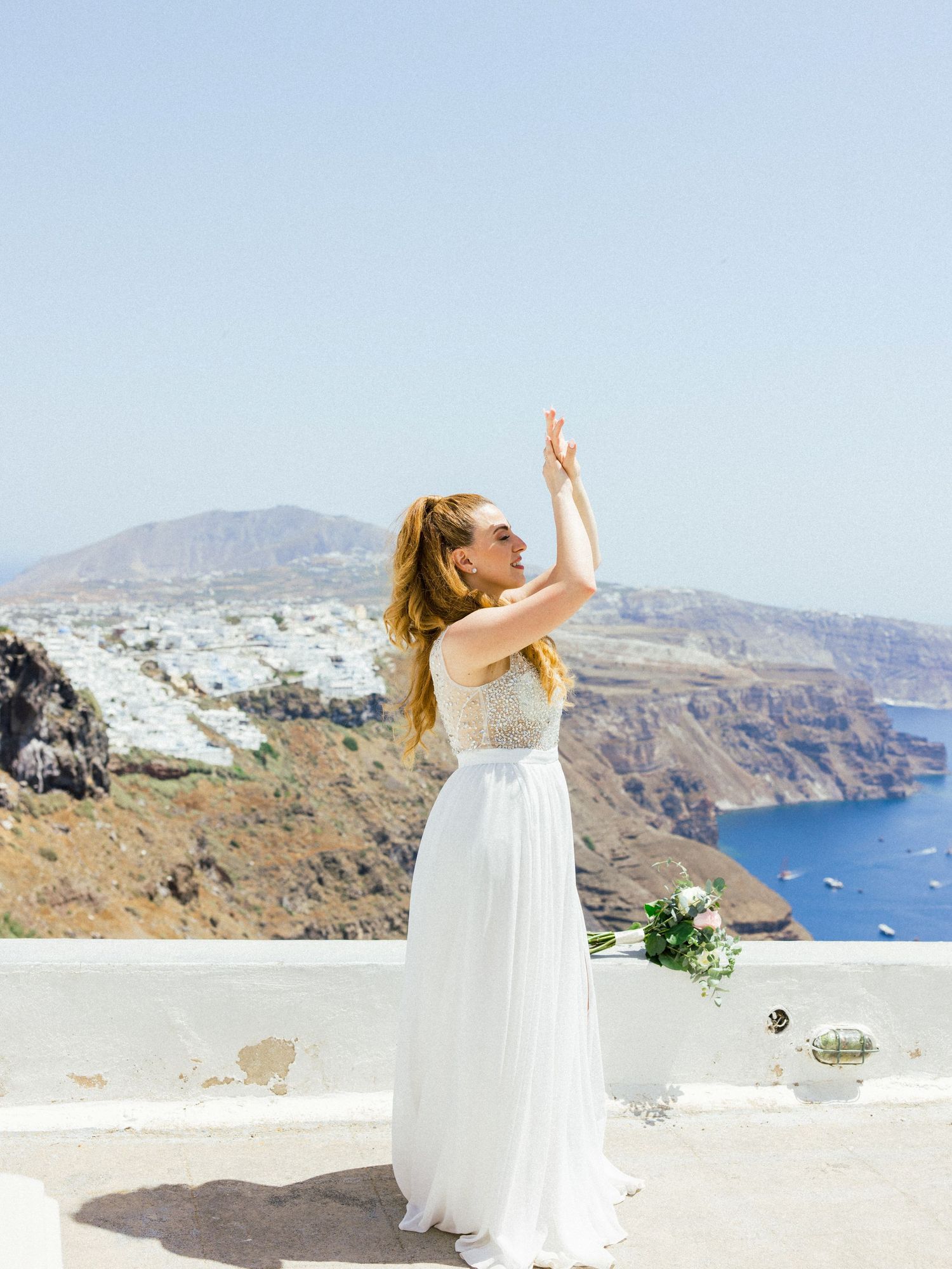 Bride in flowing white dress poses joyfully against Mediterranean coastal backdrop.