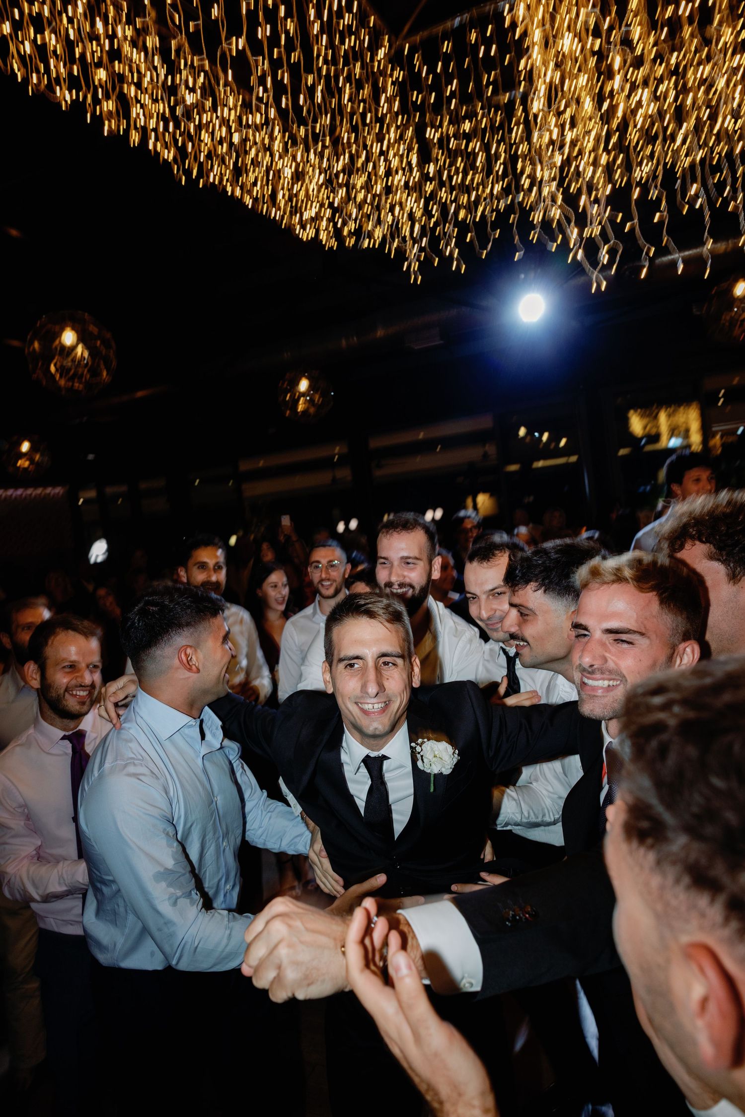 Wedding party guests laughing and celebrating under warm overhead lighting.
