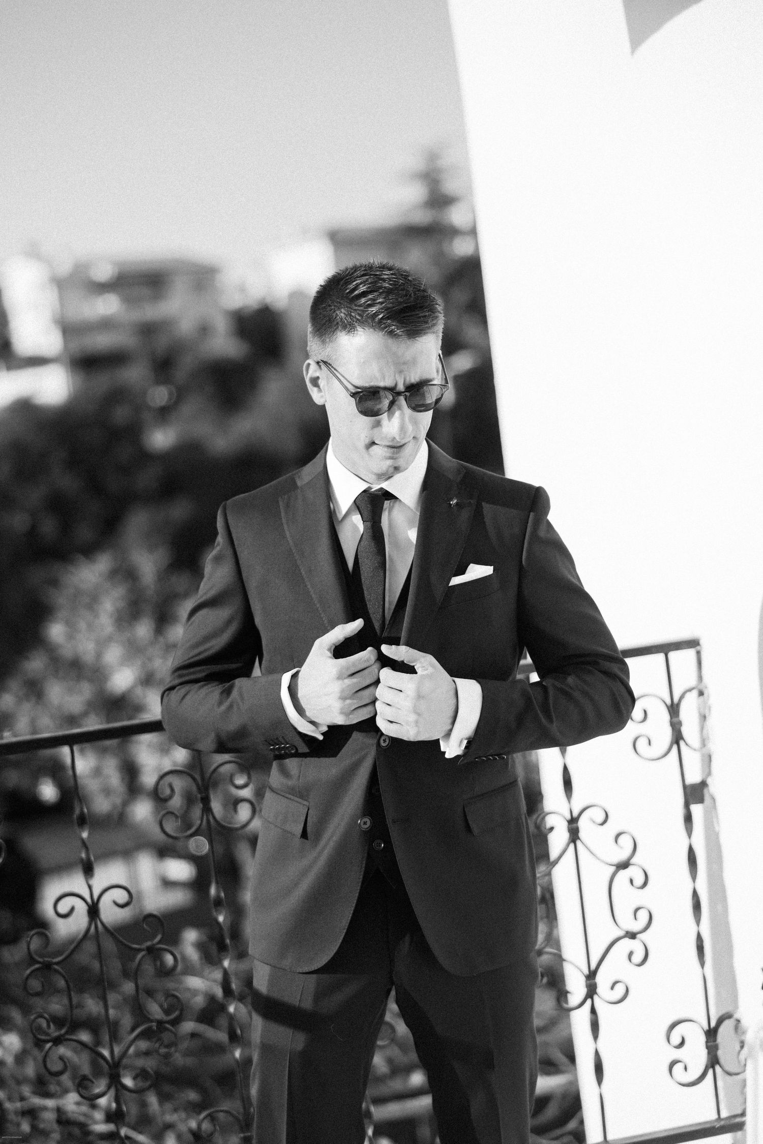 Groom in classic black suit checks his pocket square while standing on a balcony.