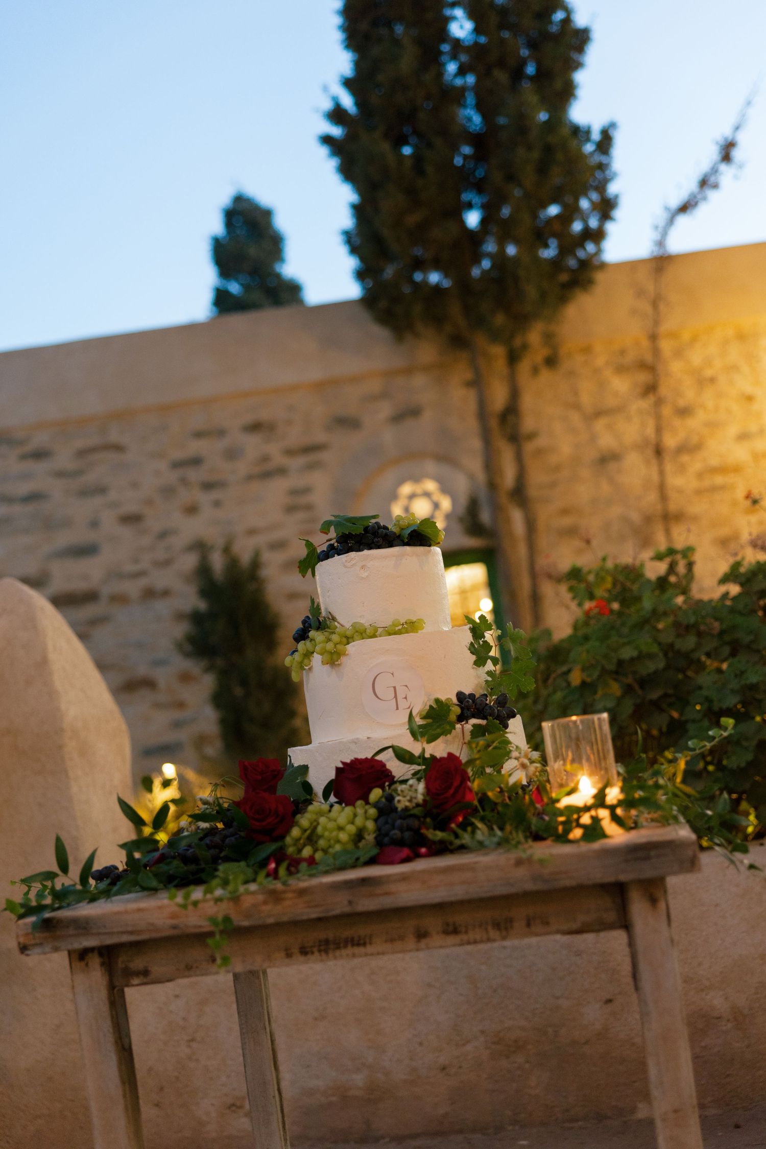 Elegant three-tier white wedding cake with monogram, adorned with greenery and red roses on rustic table at dusk.