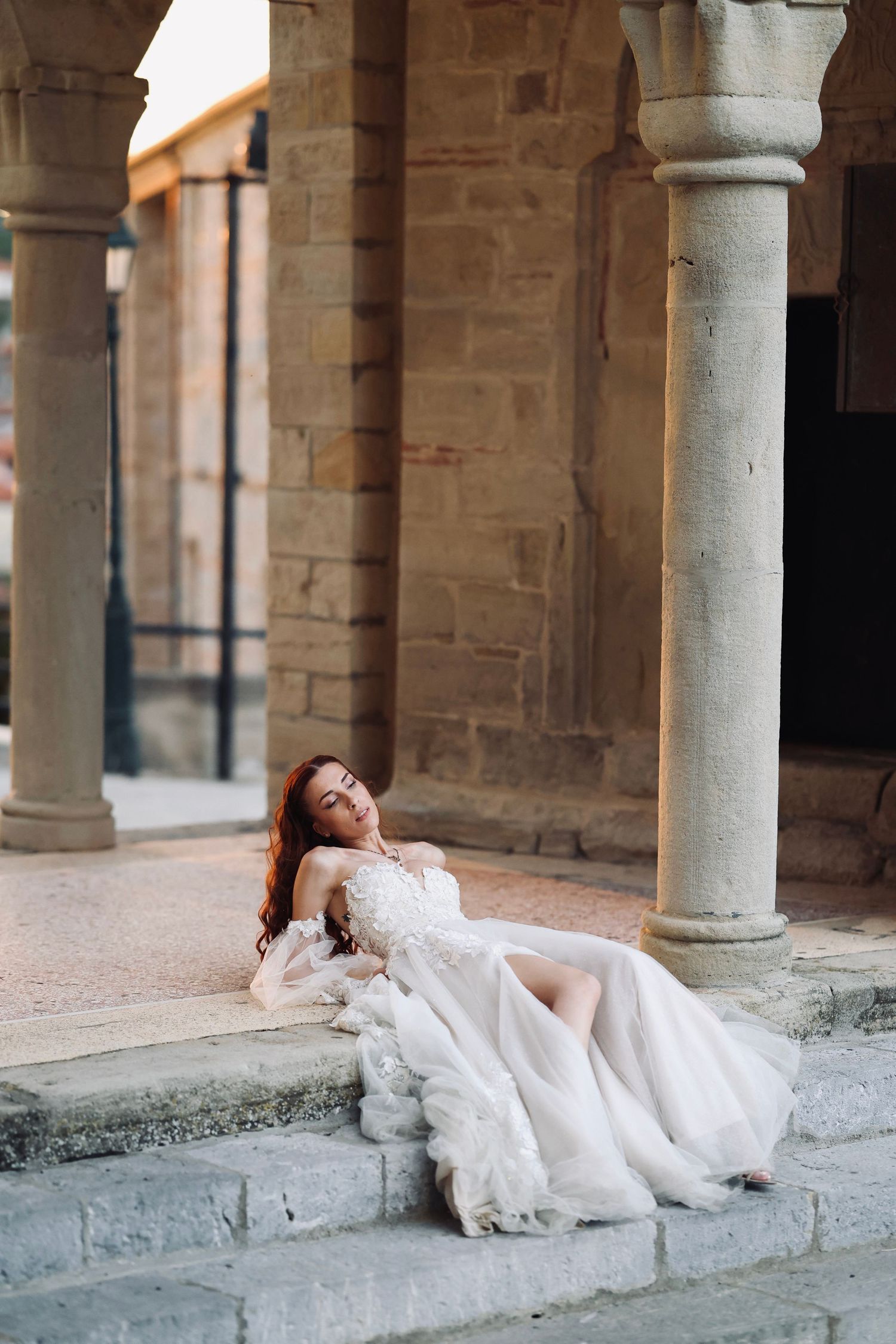 Bride in white gown resting on stone steps beside ancient columns of a historic building.