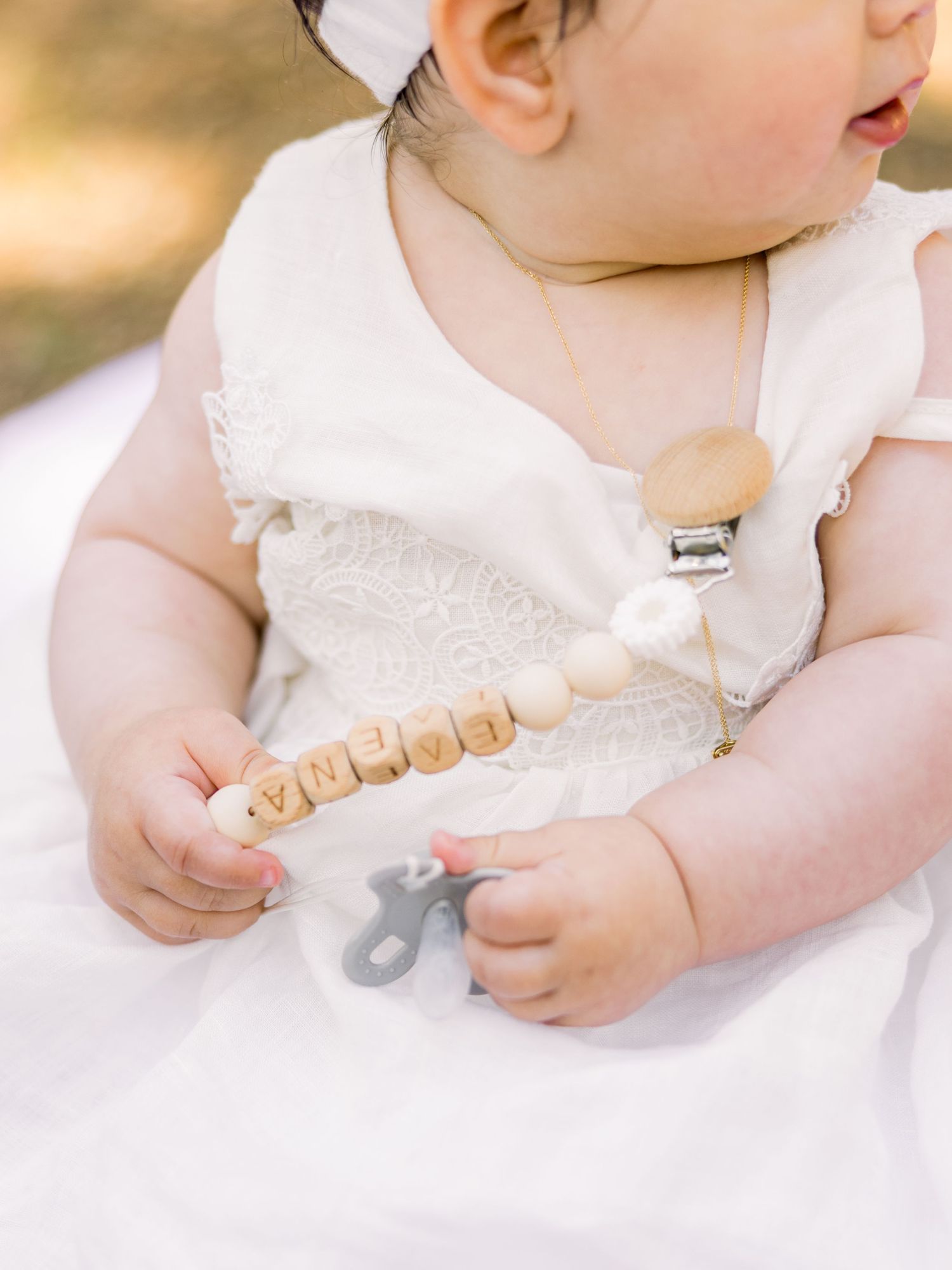 A baby in a white dress holds wooden alphabet blocks while sitting on a light colored surface.
