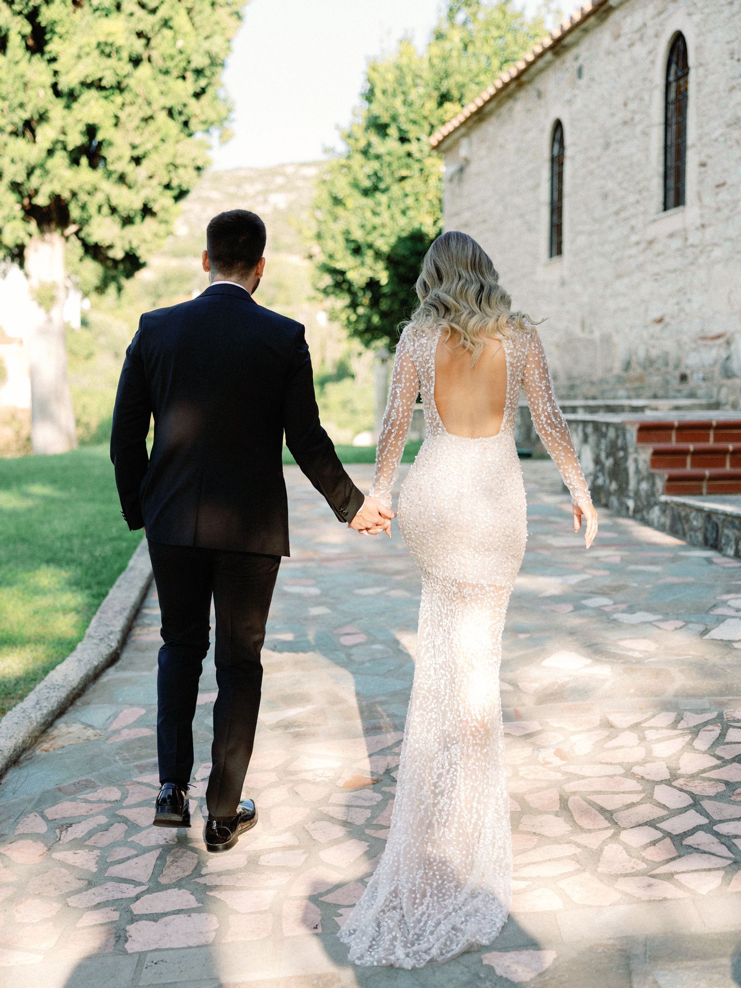 Newlyweds walk hand in hand along stone pathway by historic building in sunlit garden setting.