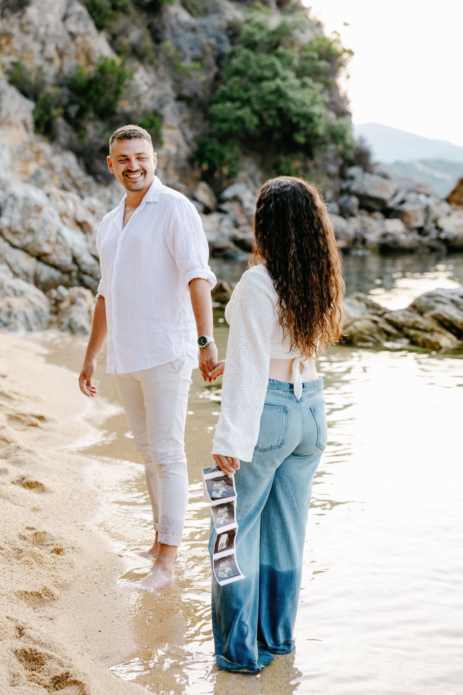 A couple walks along a rocky beach shore wearing coordinated white and blue outfits.
