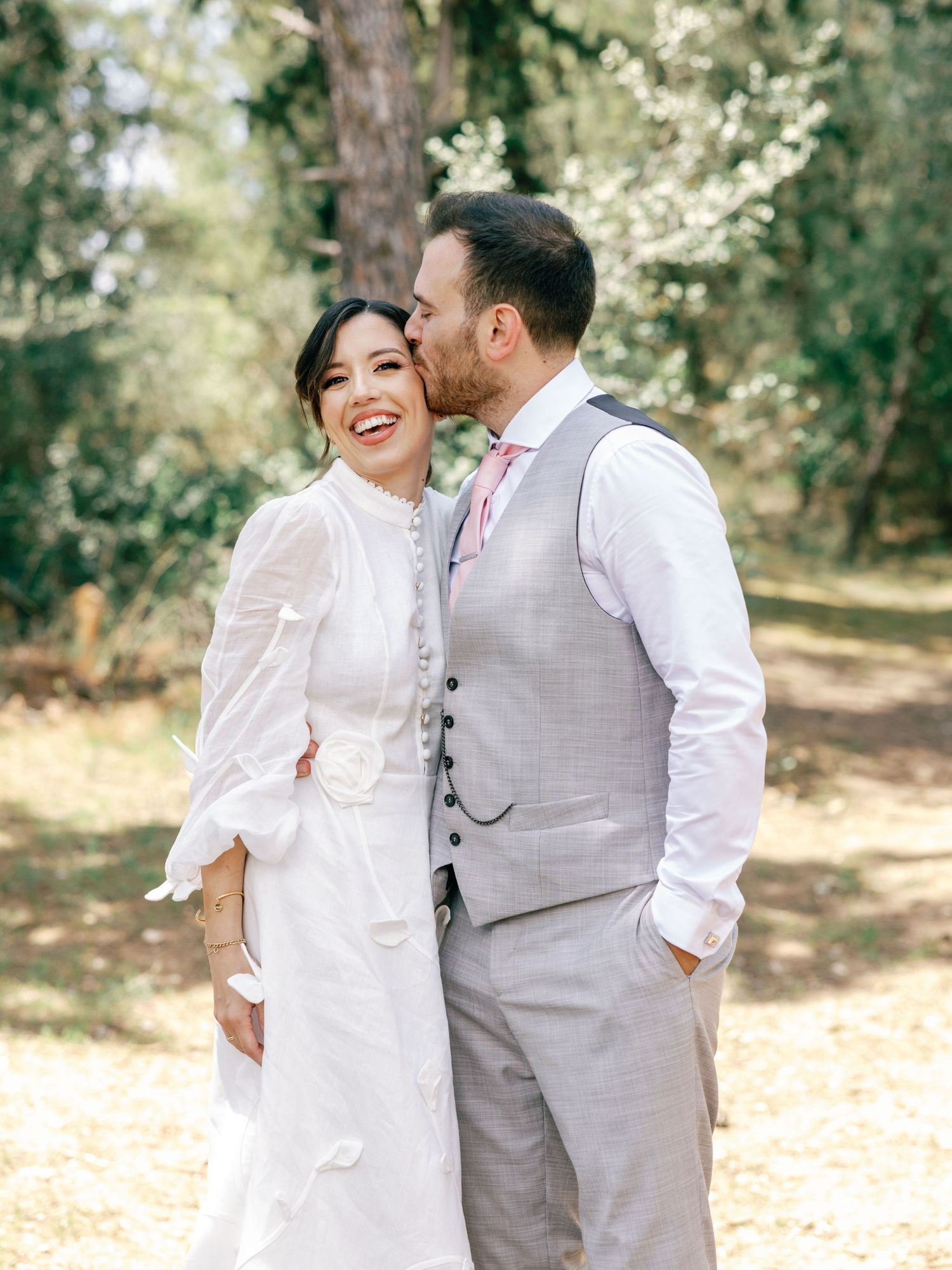 A couple shares joyful moments in a sun-dappled outdoor setting wearing light-colored wedding attire.