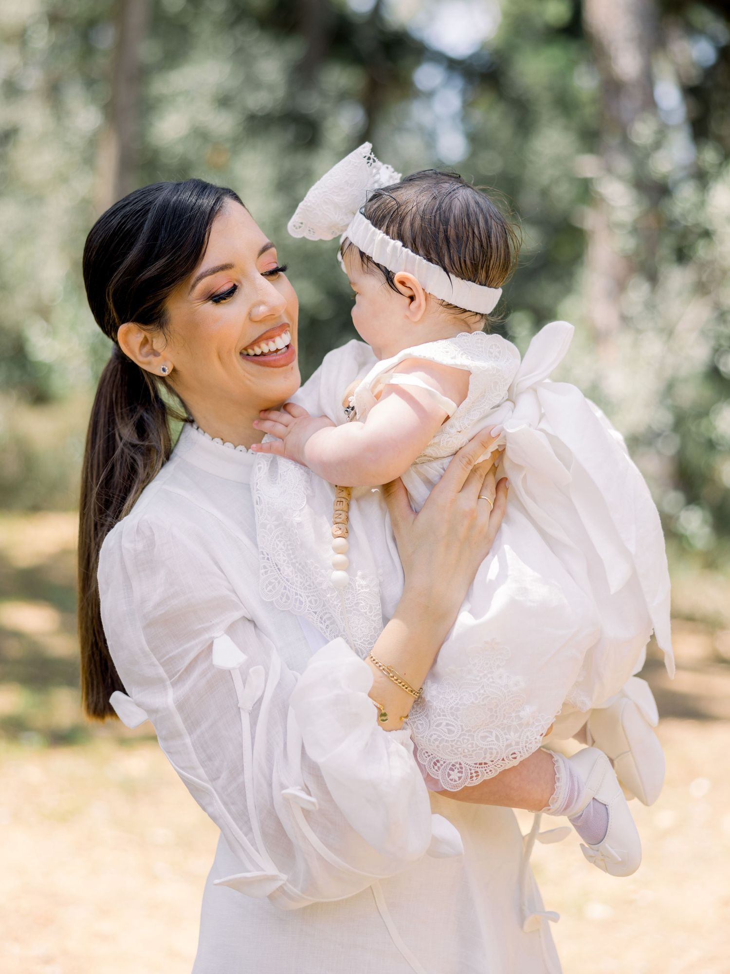 A tender moment captured outdoors as a woman in white holds a baby wearing a bow headband in a sunlit forest setting.