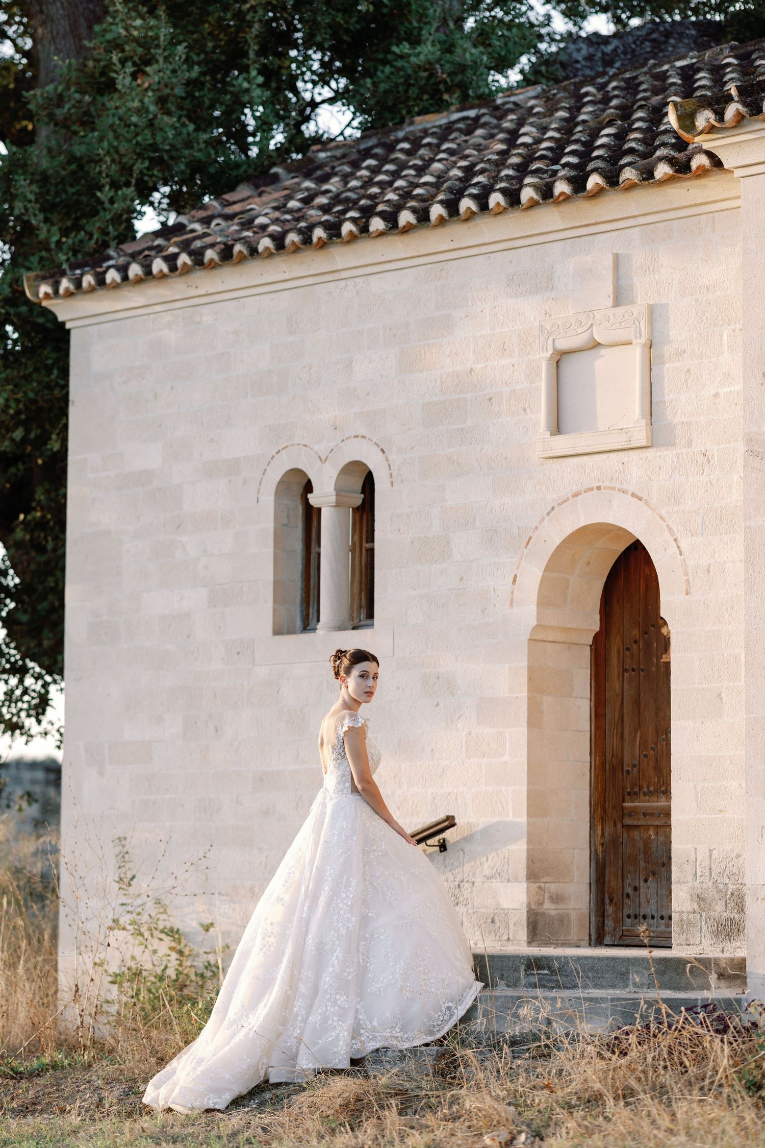 A bride in a flowing white dress poses against a rustic white stone chapel wall.
