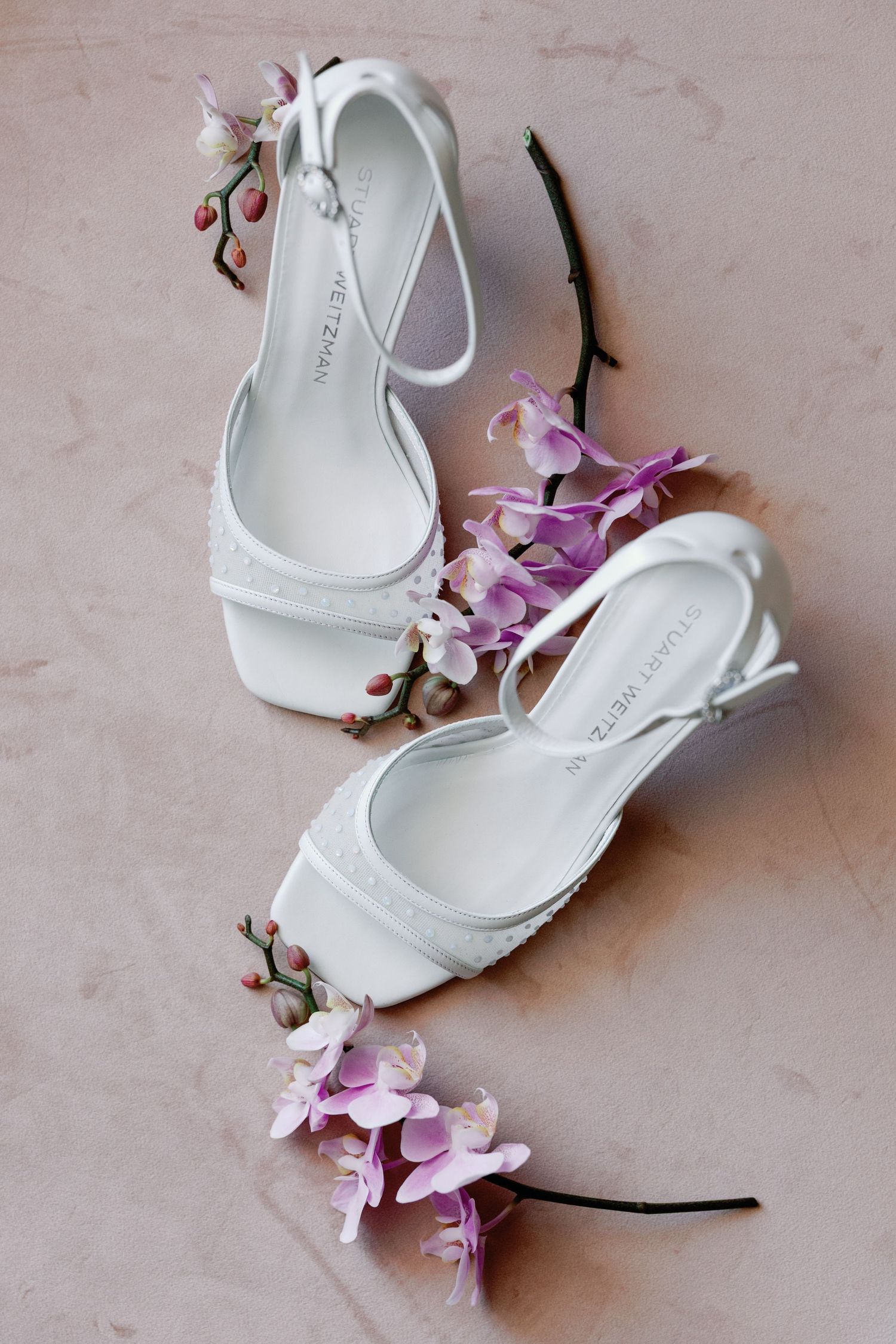 White bridal shoes with delicate pink orchid flowers arranged on a beige surface.