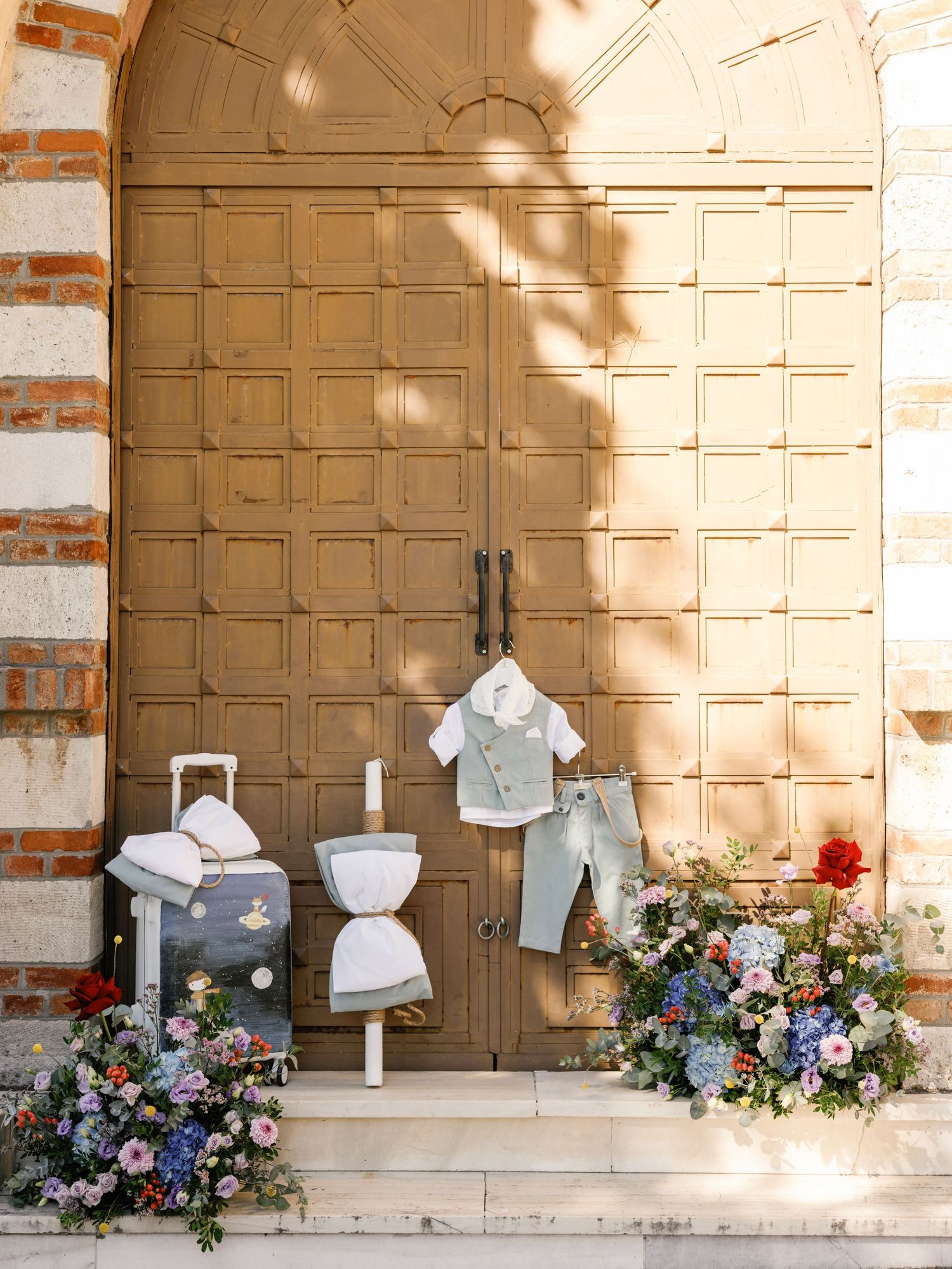 Small formal outfits and floral arrangements decorate an elegant wooden door entrance with sunlight casting soft shadows.