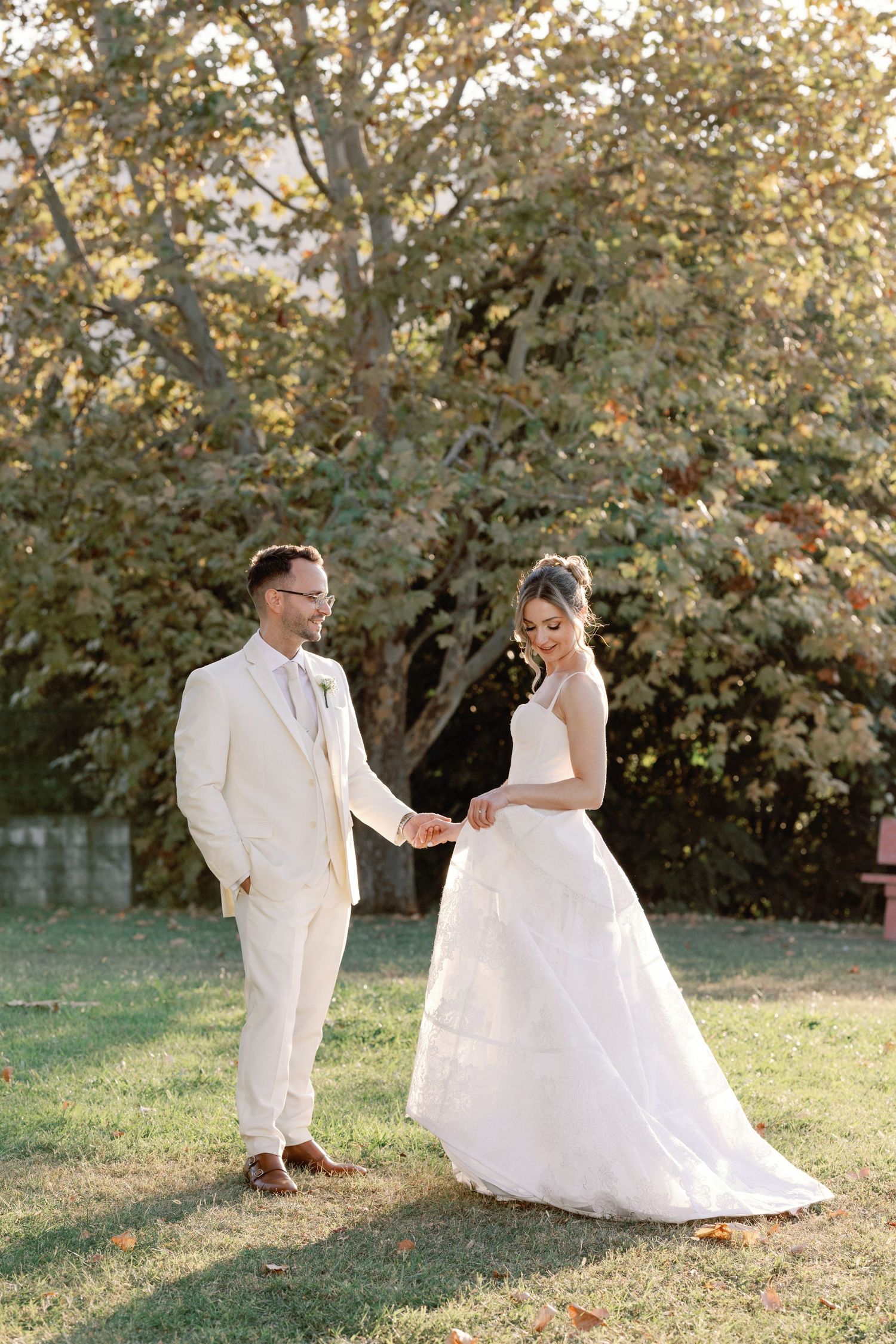 Newlyweds in white attire pose together in front of blooming trees during golden hour.