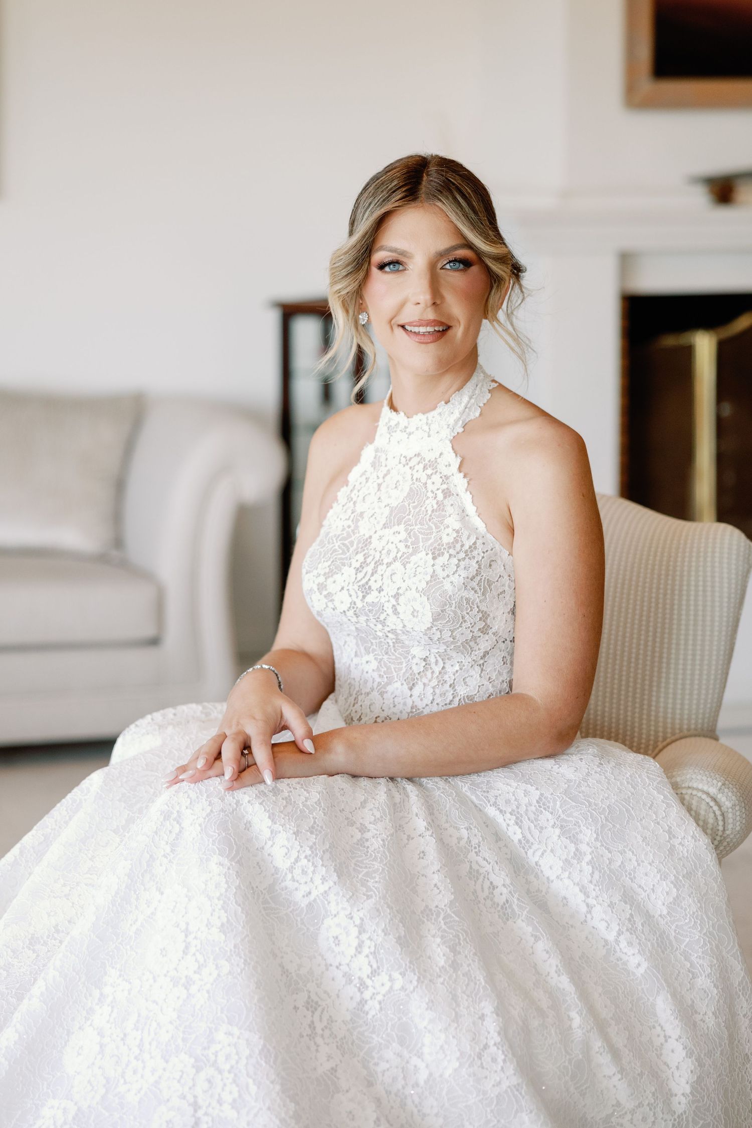 Bride in luxurious white lace dress sitting elegantly in a bright interior setting.