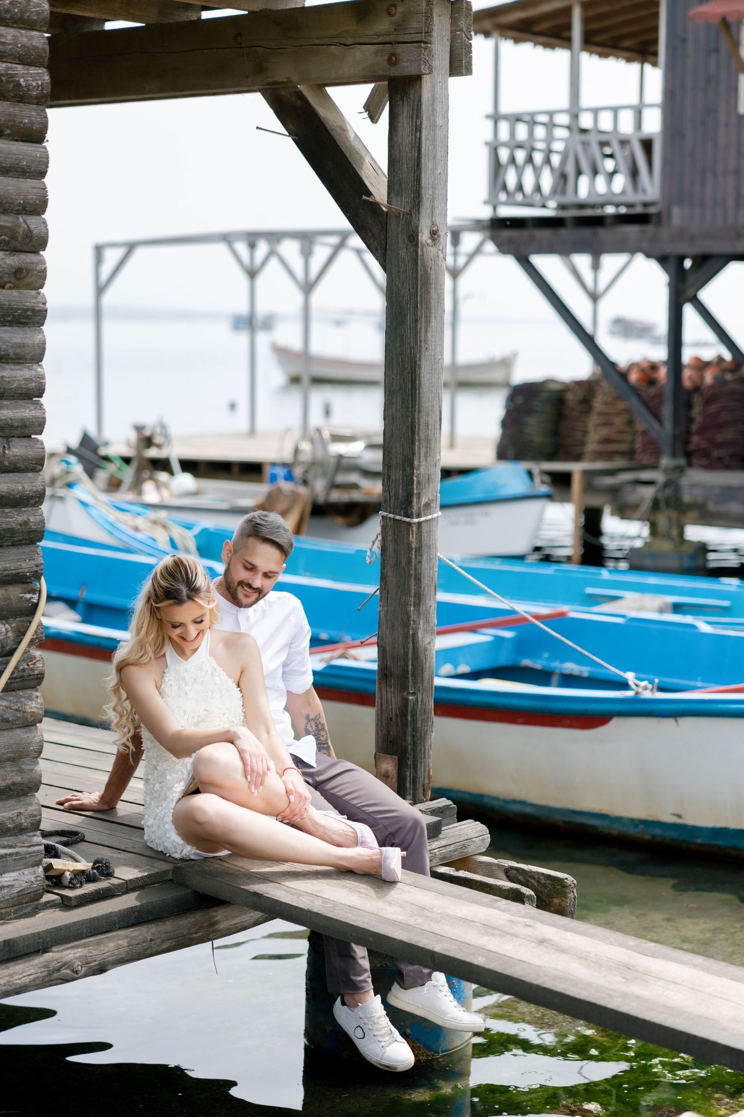 Couple sitting together on a wooden dock by colorful boats, sharing a romantic moment at a seaside marina.