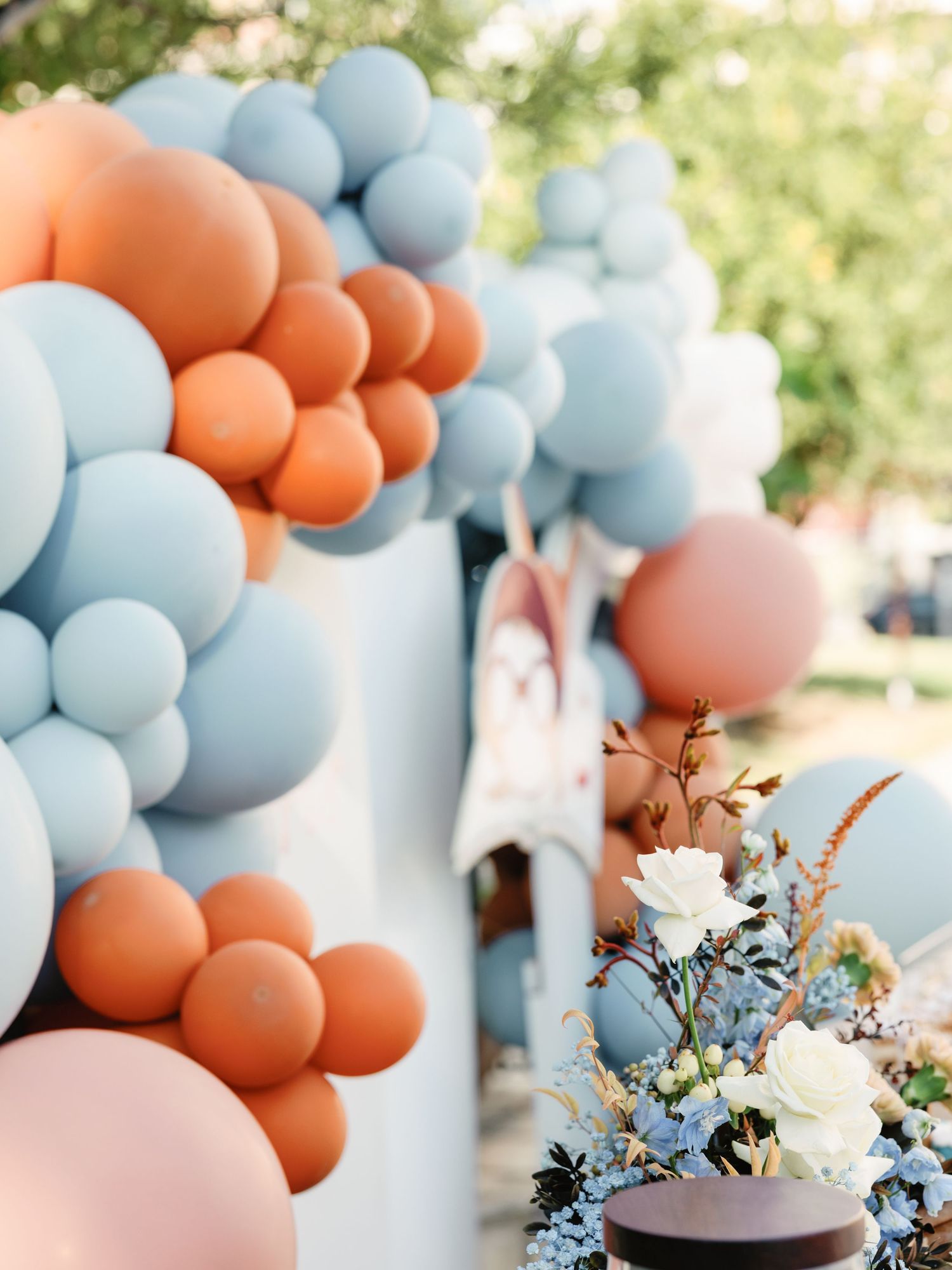 Light blue and peach balloon garland with white floral arrangements creates an elegant party decoration backdrop.