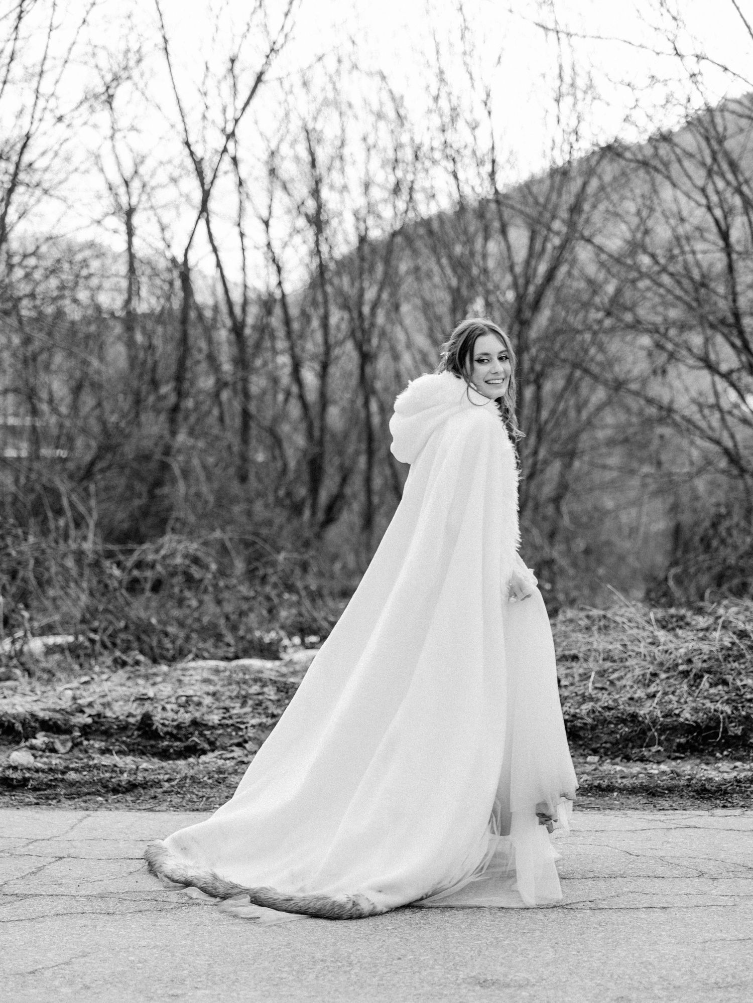 A dramatic black and white bridal portrait featuring a flowing cape against a winter landscape.