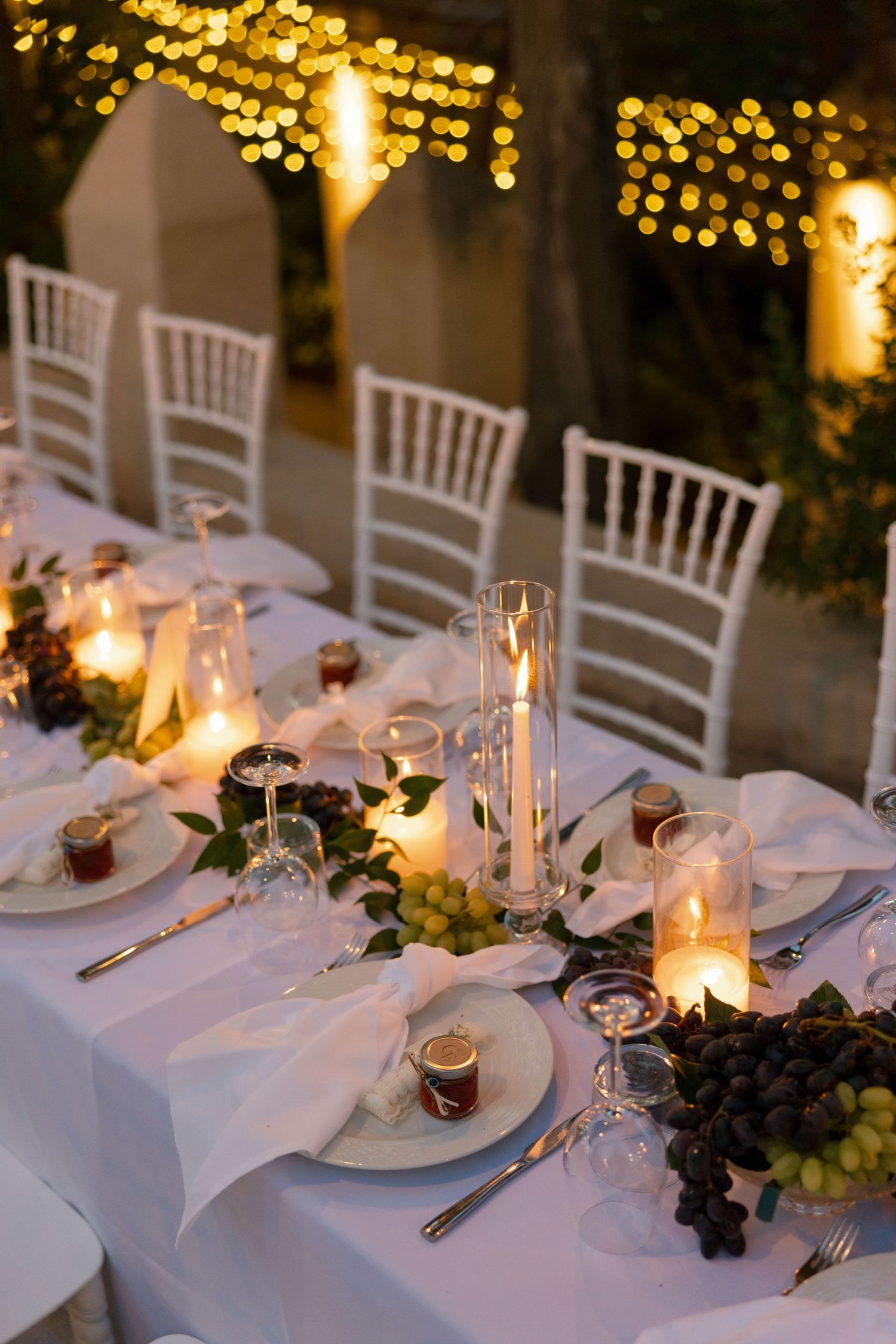Elegant outdoor wedding reception table with candles, grapes, and white Chiavari chairs under warm string lights at dusk.