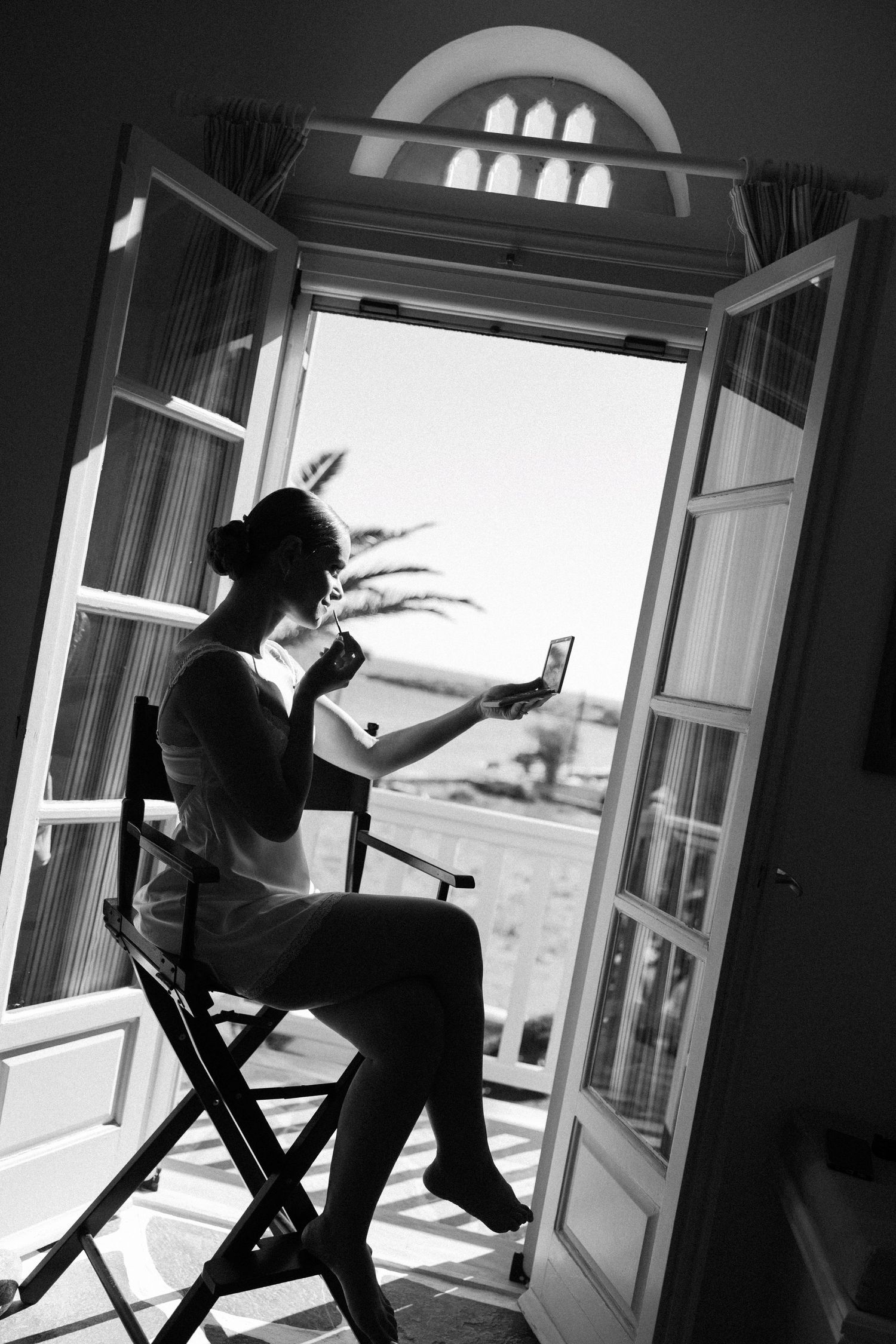 Black and white photo of a woman sitting in a director's chair by open French doors, applying makeup with a mirror.
