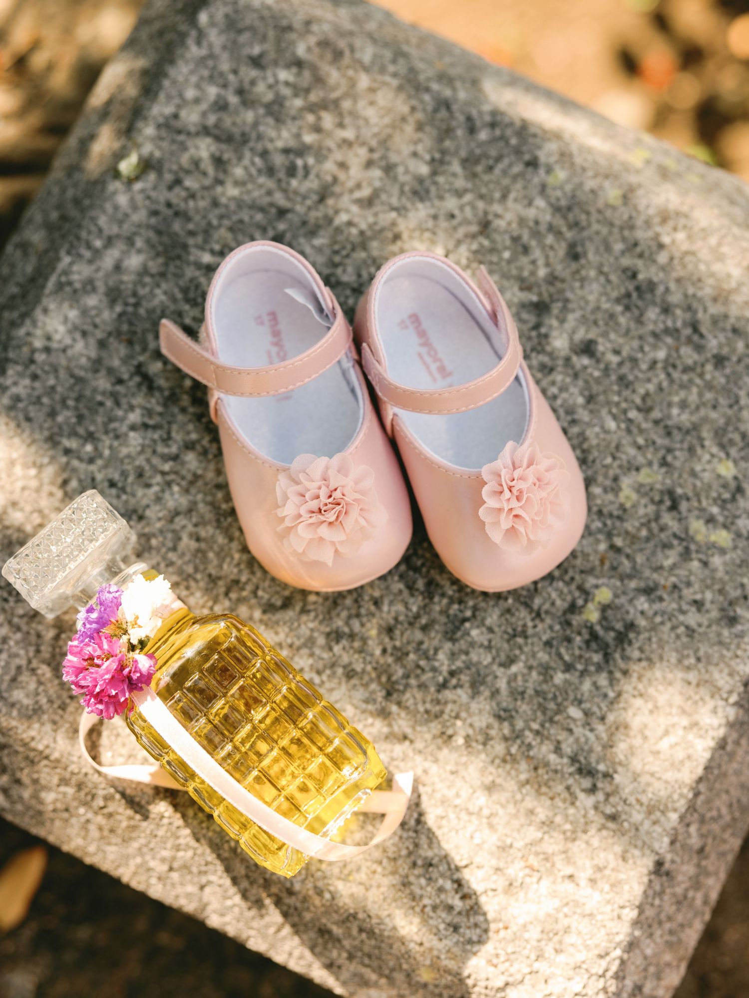 Small pink baby shoes and a perfume bottle arranged on a stone surface in a decorative still life composition.