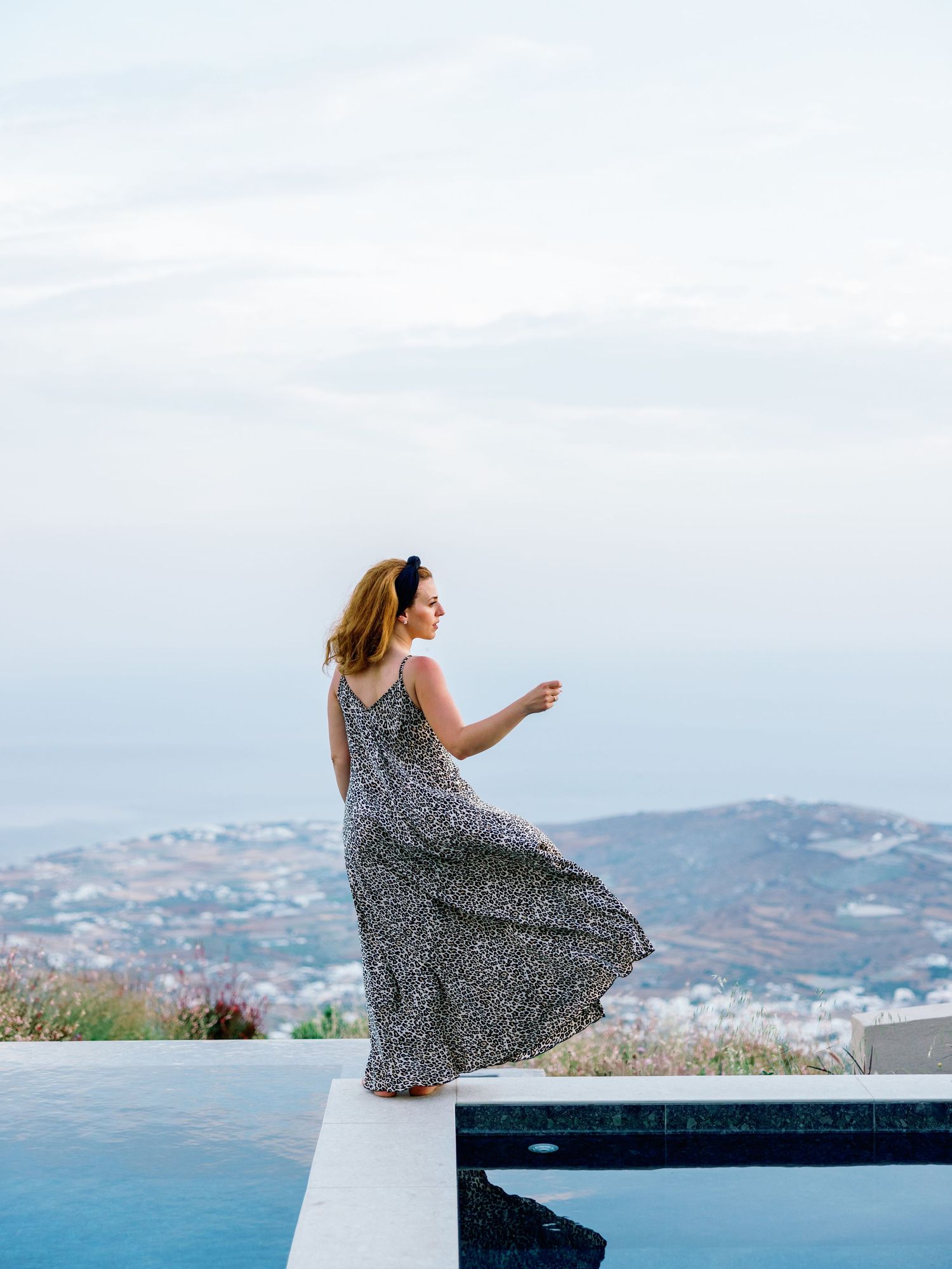A person in a flowing dress sits on a white wall overlooking the Mediterranean coastline.