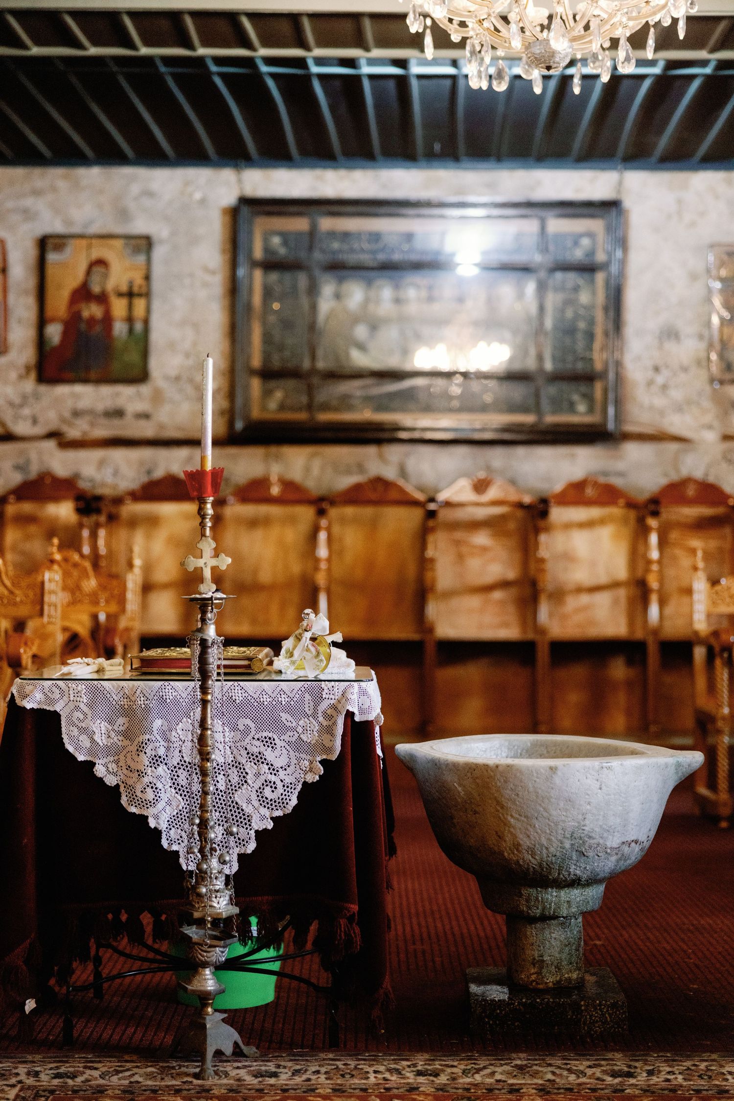 Church interior with ornate baptismal font, lace altar cloth, and religious icons on stone walls.