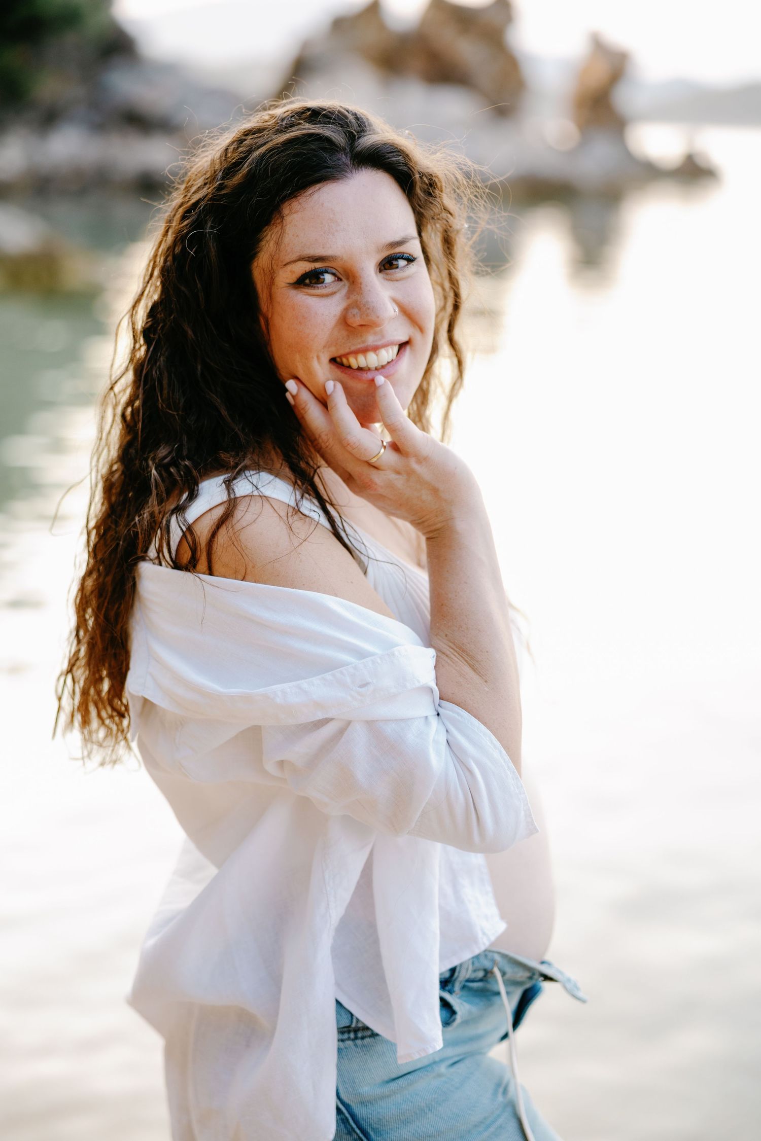A woman in white shirt and jeans laughs joyfully during a beach photoshoot.