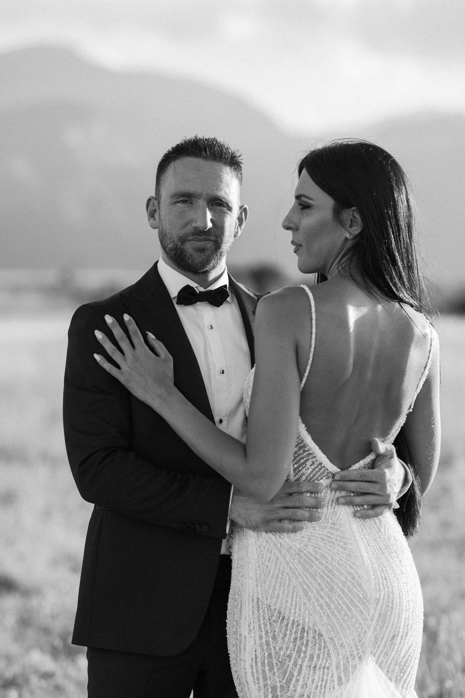 A couple embraces in formal wedding attire against a mountain landscape.