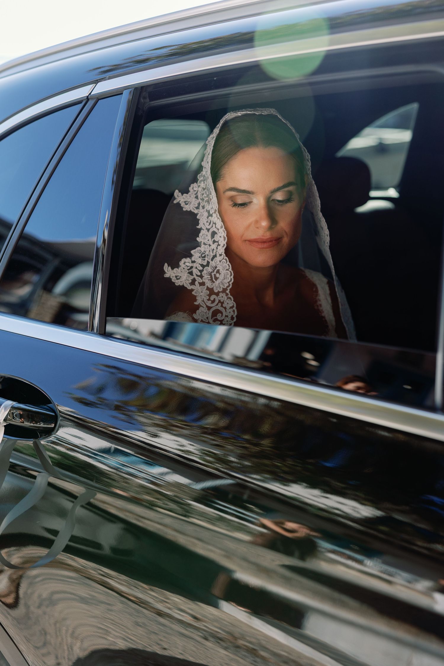 Elegant bride wearing a lace veil sits in a luxury black car, looking down gracefully on her wedding day.