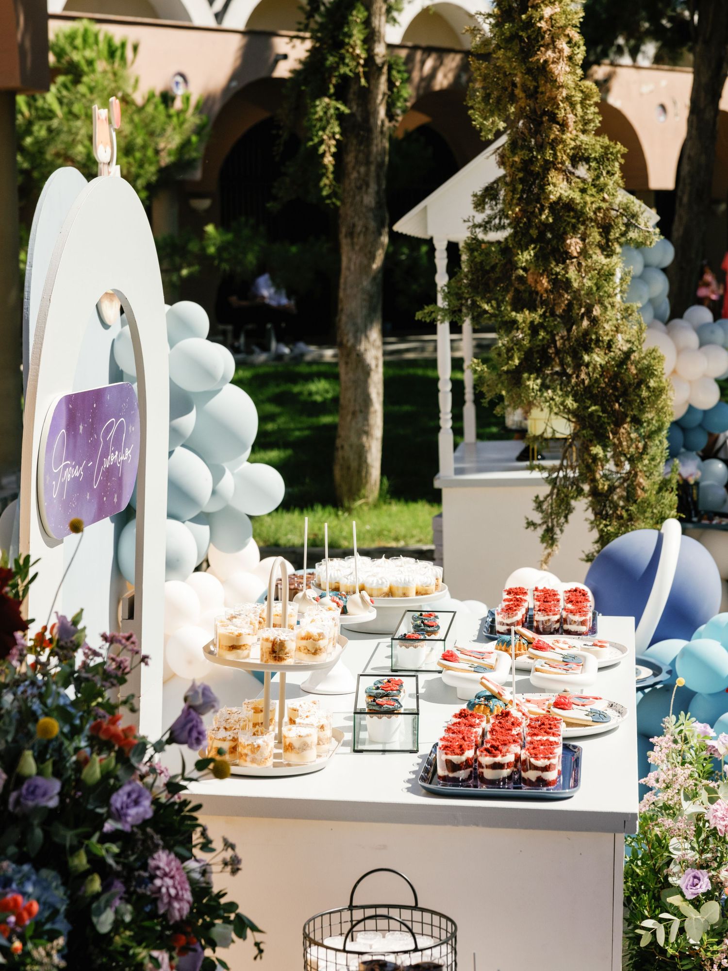 Elegant outdoor dessert table with tiered displays of pastries and treats decorated with blue balloon garlands.