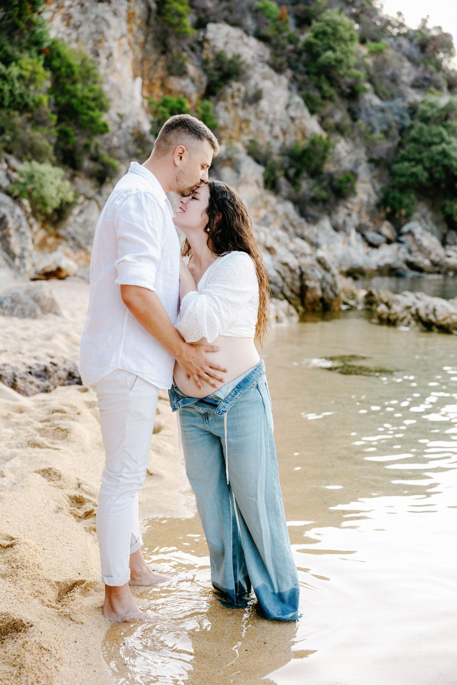 Romantic beach maternity photos with couple standing in shallow water near rocky cliffs.
