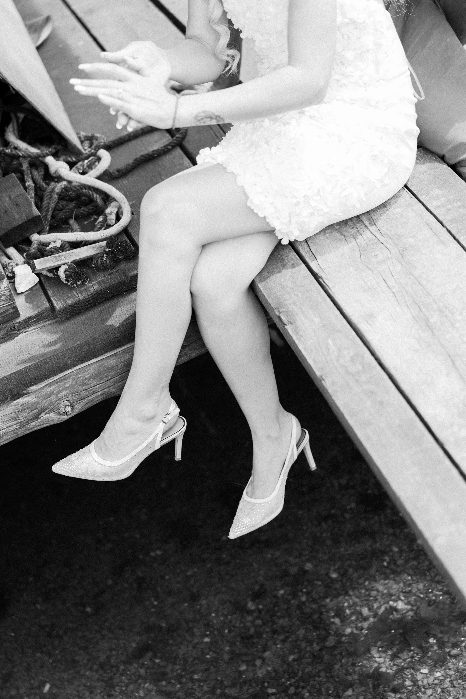 Black and white photo of a bride in white dress and heels sitting on wooden dock steps.
