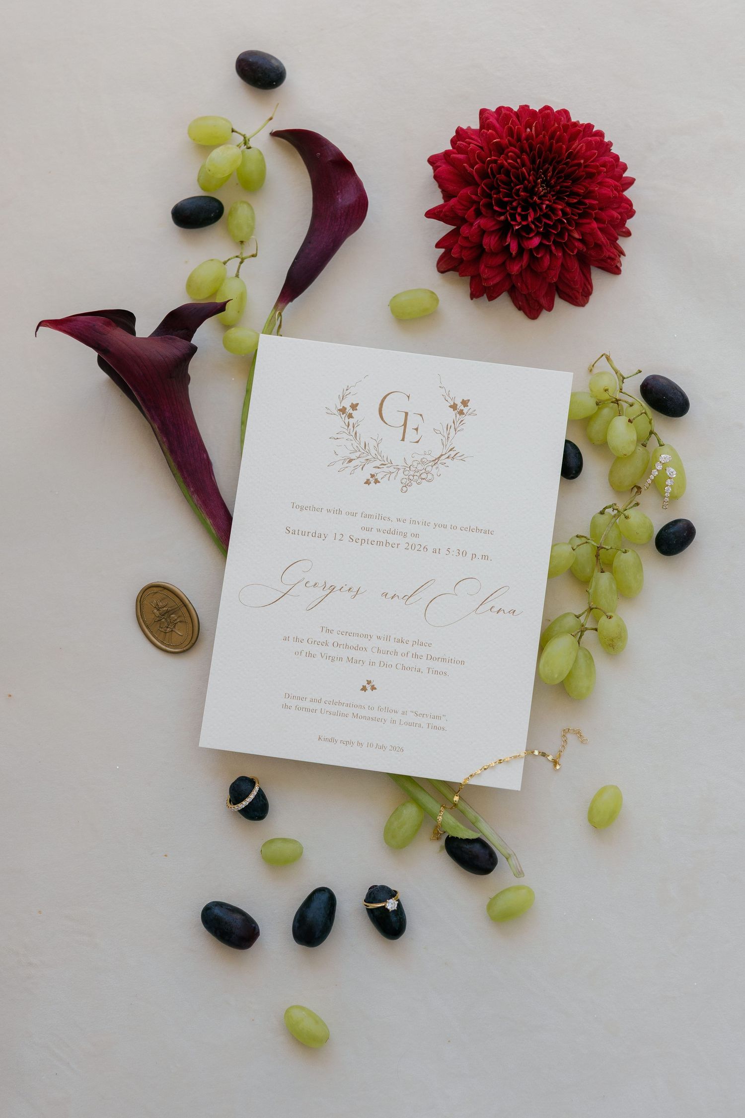Elegant wedding invitation surrounded by green grapes, dark olives, red dahlia, and purple calla lilies on white background.