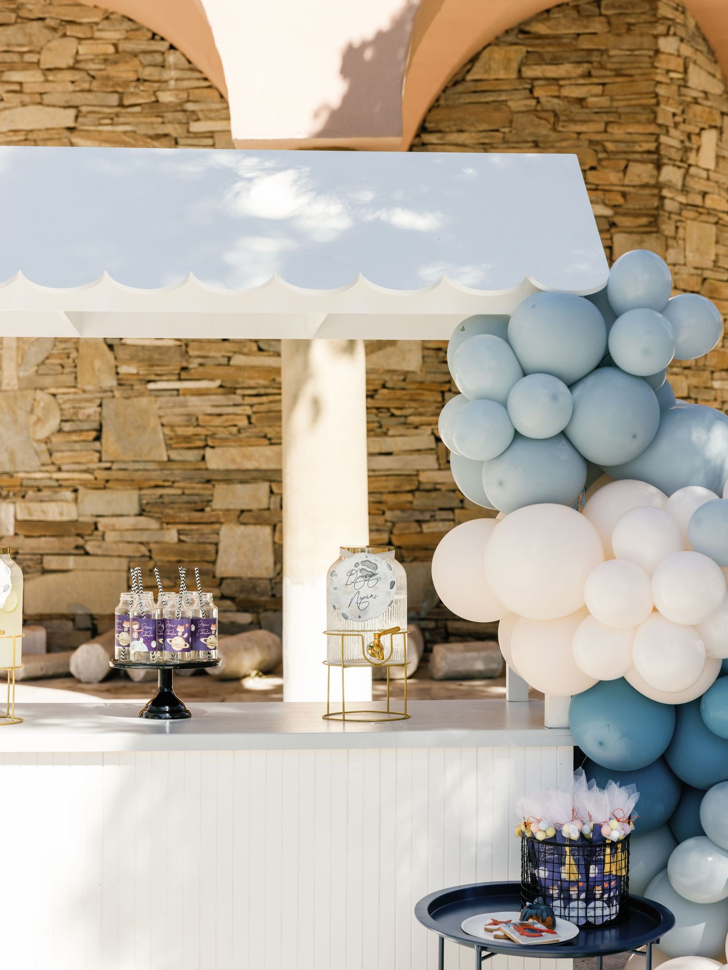 Elegant party setup with blue and white balloon arch, stone archways, and dessert display table with lanterns and cakes.