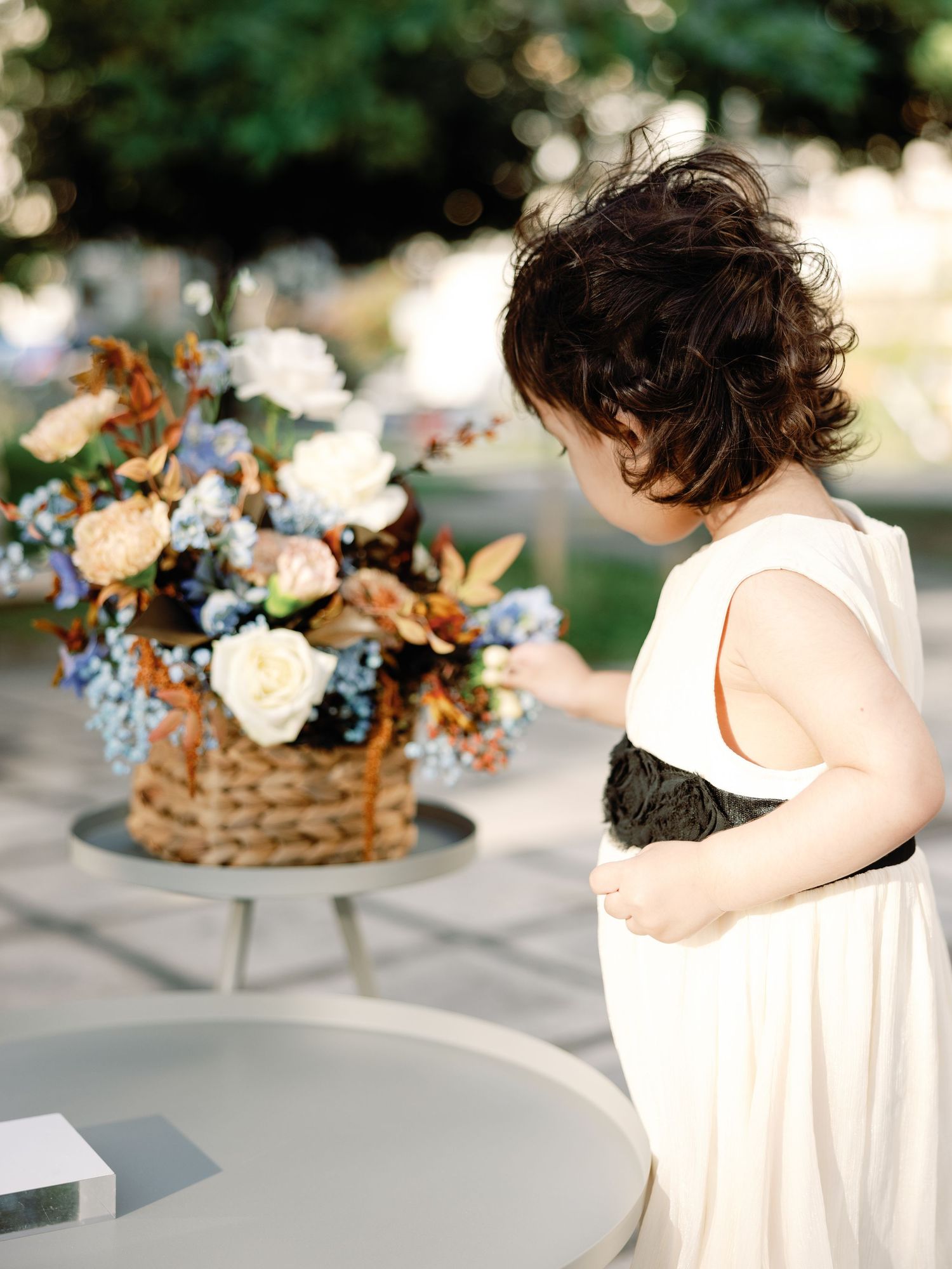 Elegant floral centerpiece with white roses and blue accents sits beside a figure in a flowing white dress at an outdoor event.
