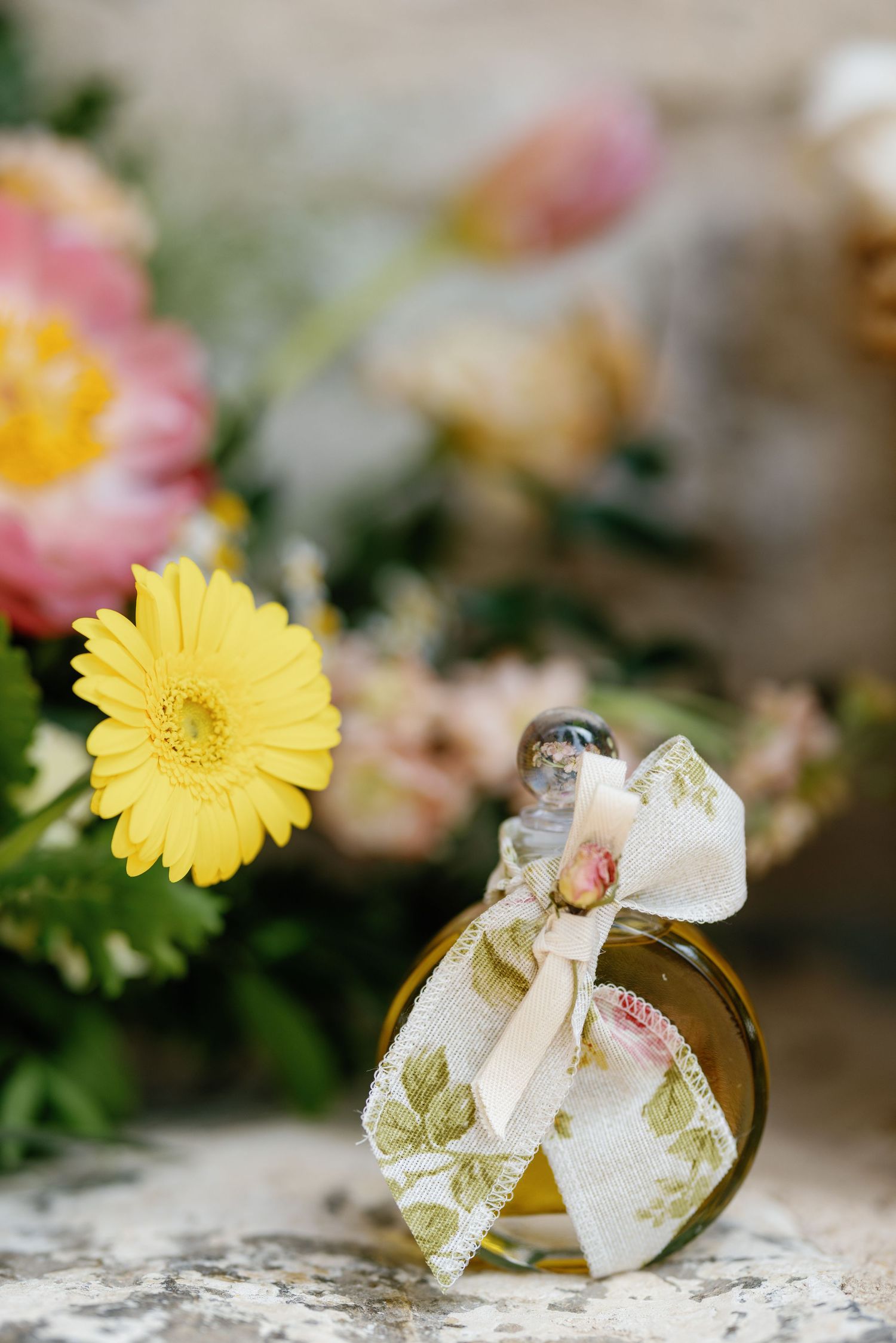 A decorative perfume bottle with floral ribbon sits among yellow and pink flowers.